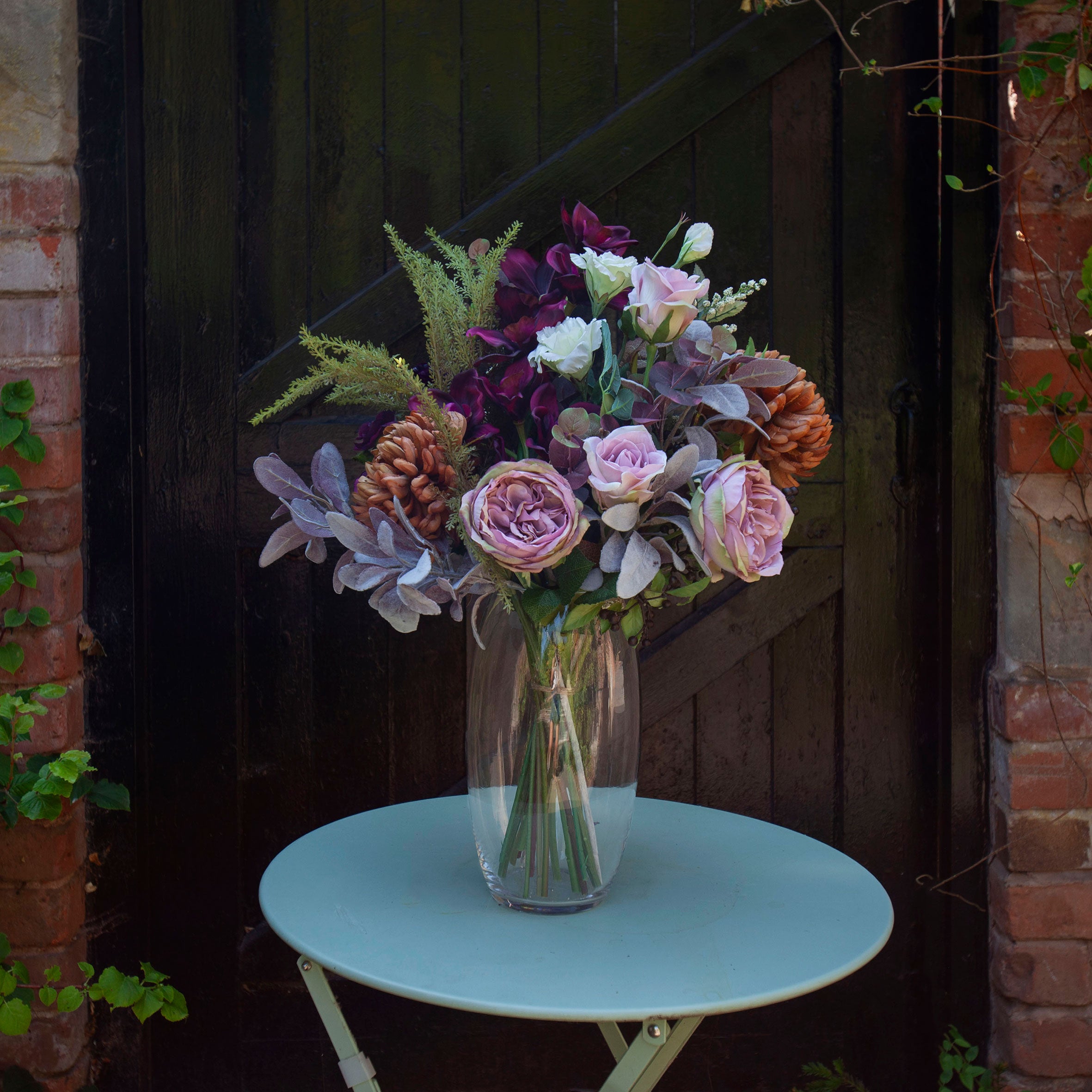Artificial garden roses, burgundy orchids and bronze chrysanthemums with silvery foliage, arranged in a tall clear glass vase against a rustic dark wooden backdrop.