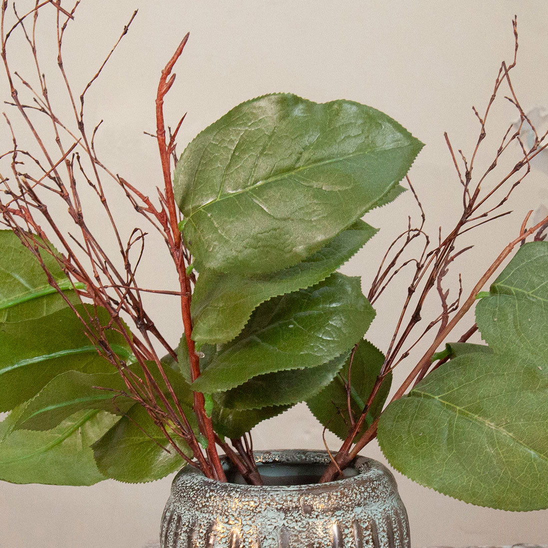 Close up of artificial green salal leaf stems with delicate brown branches displayed in a textured metallic vase