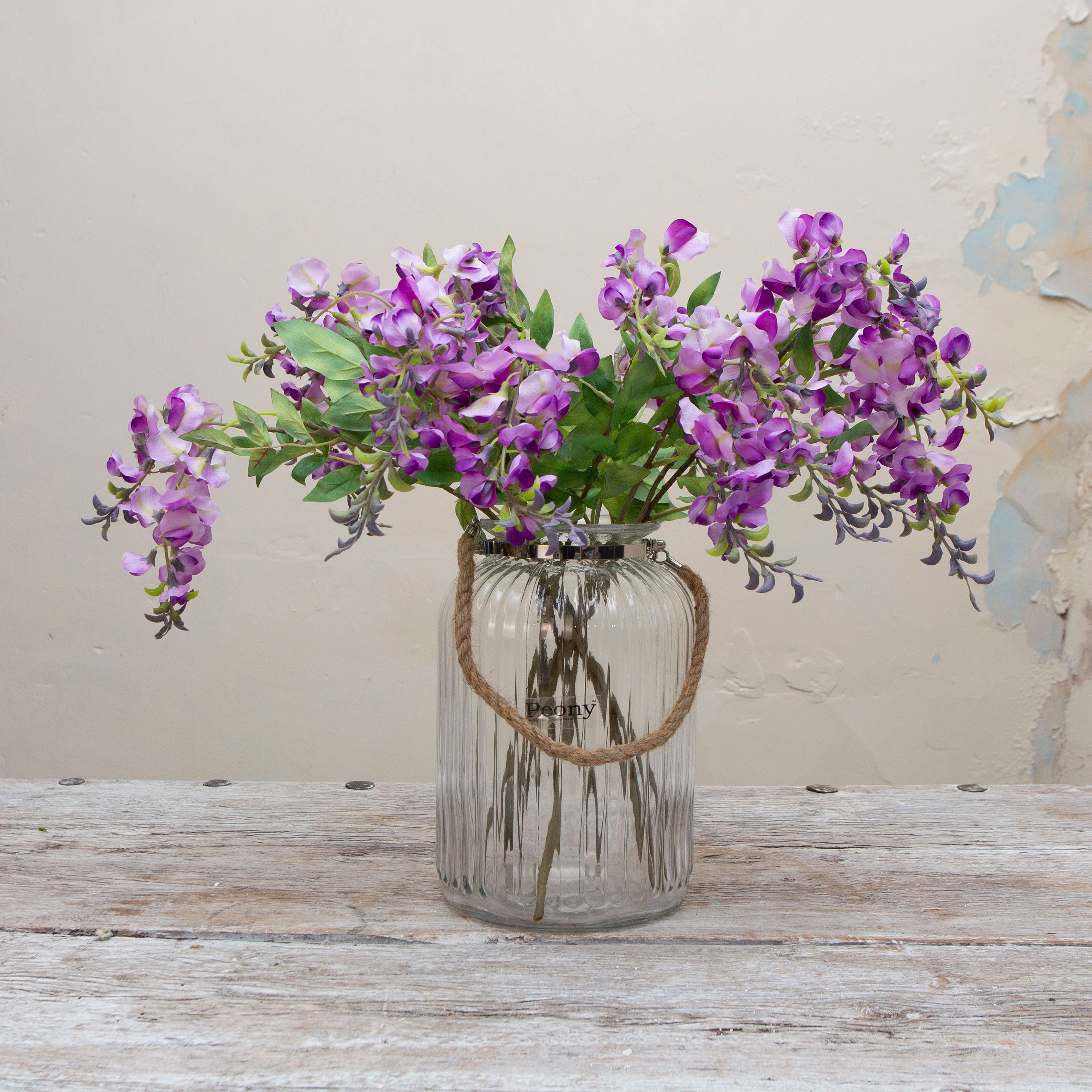 Artificial purple wisteria stem displayed in a vase, showcasing trailing clusters of delicate purple flowers and natural green leaves