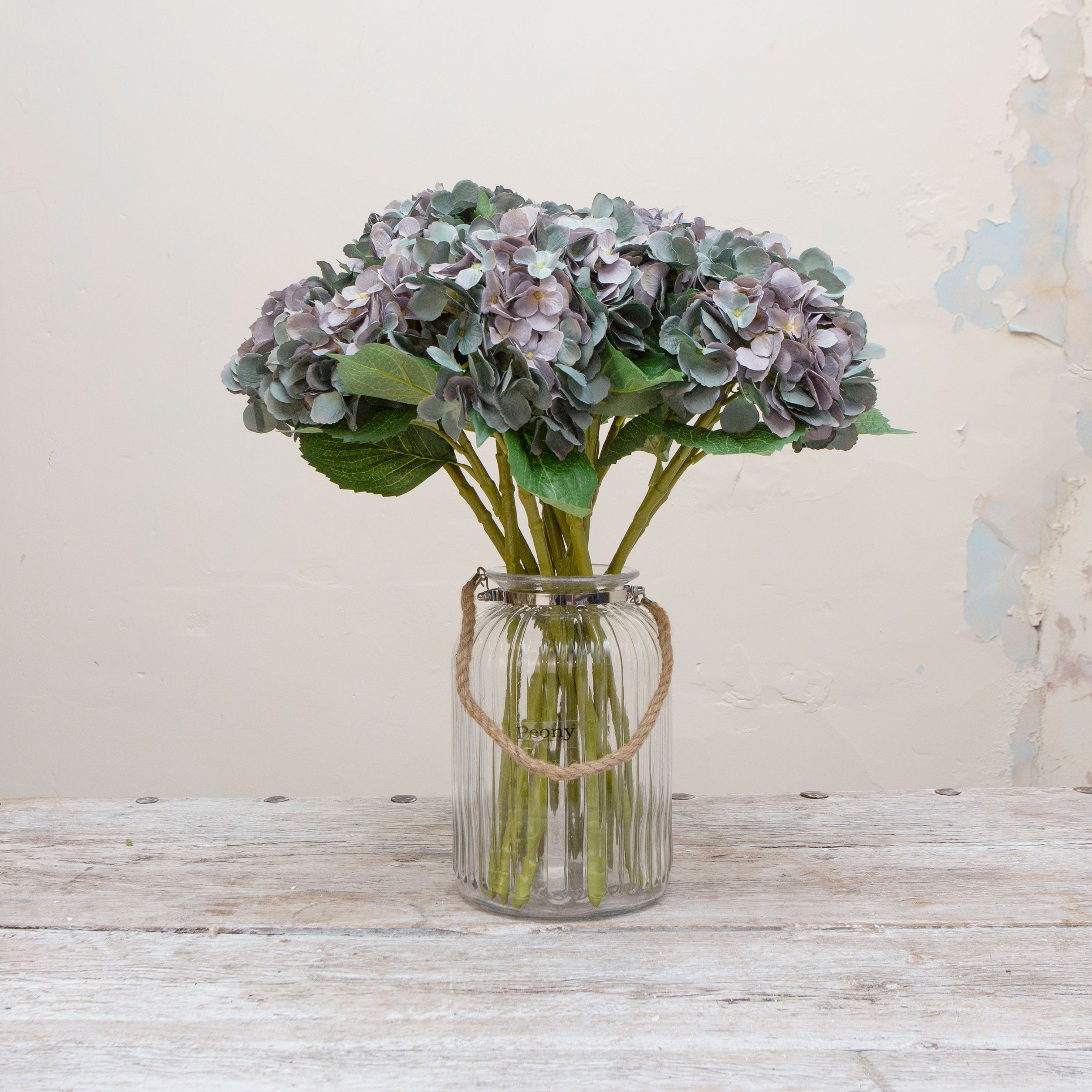 Artificial hydrangea stems with soft blue-purple flower heads and green leaves, displayed together in a glass vase for presentation
