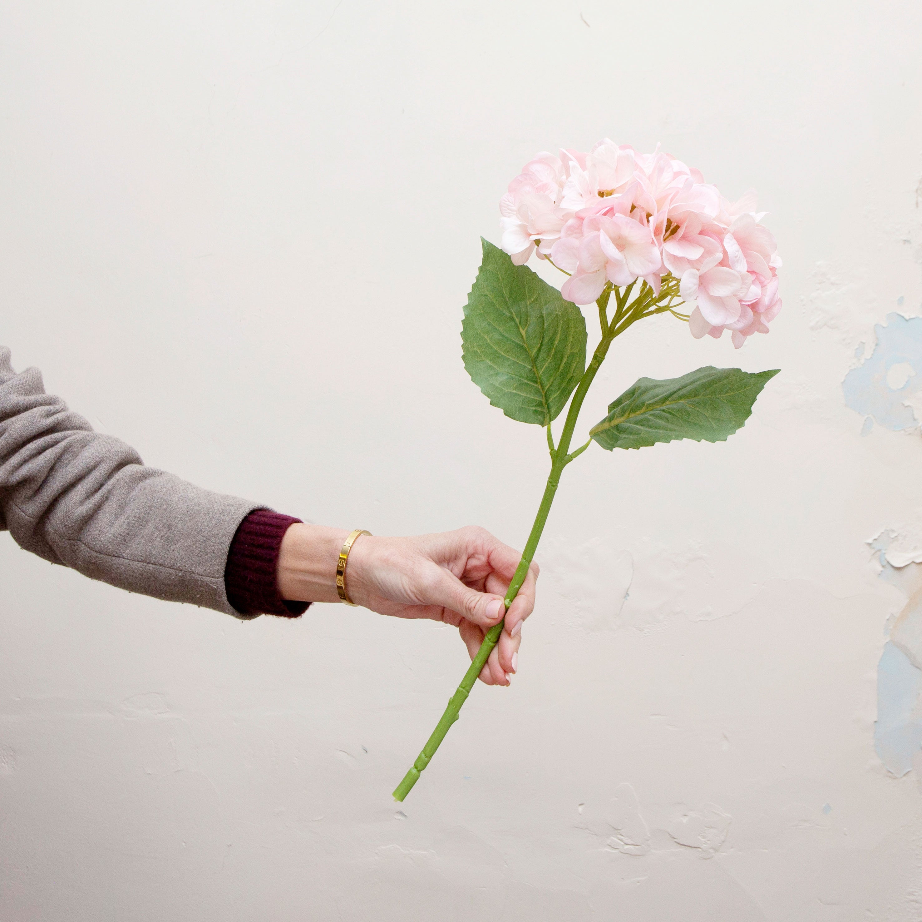 Artificial pale pink hydrangea single stem being held in hand, featuring a soft rounded flower head with blush pink petals and realistic green leaves on a sturdy stem.