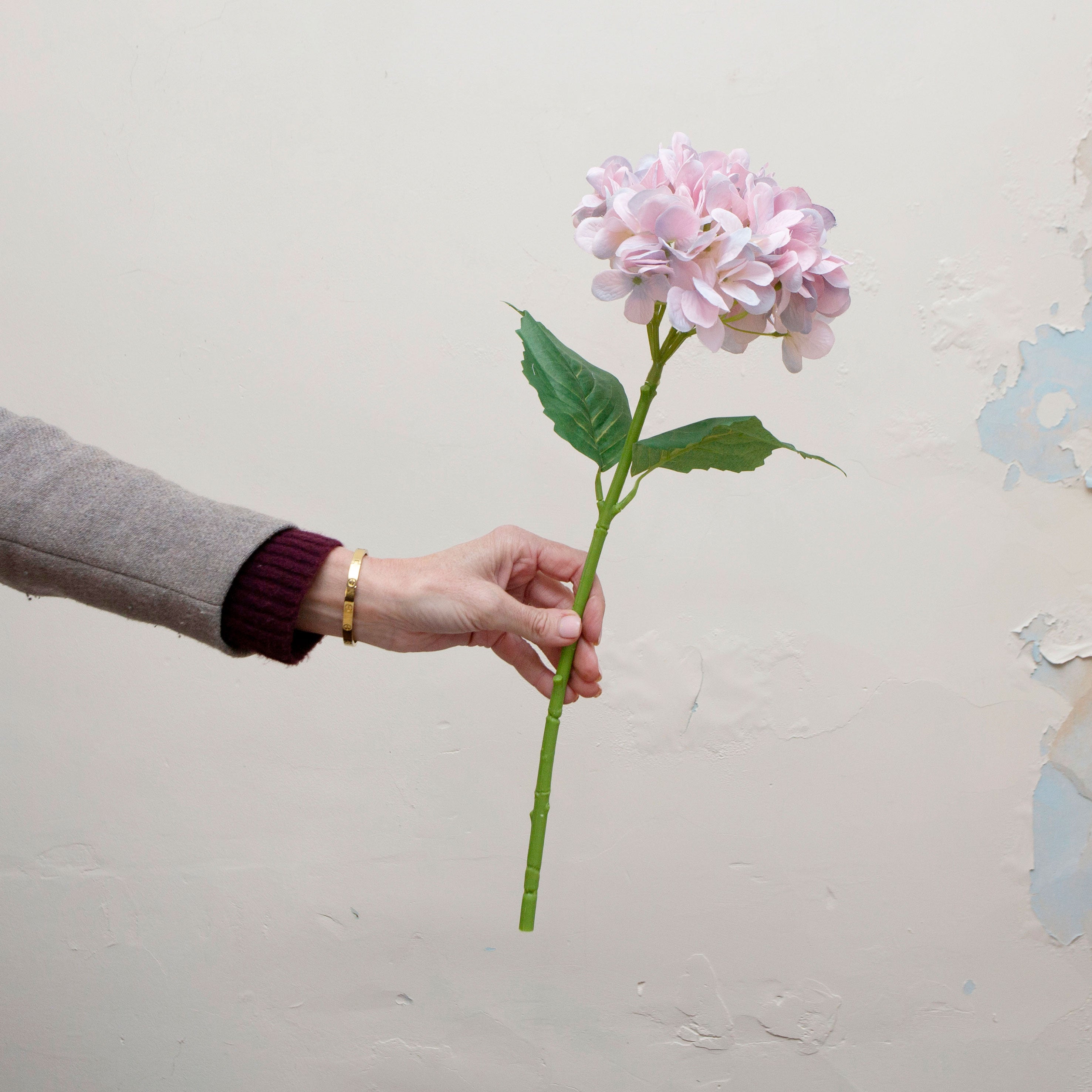 Artificial lilac-pink hydrangea single stem being held in hand, featuring a rounded cluster of soft pastel petals with subtle lilac and blush tones, paired with realistic green leaves on a sturdy stem