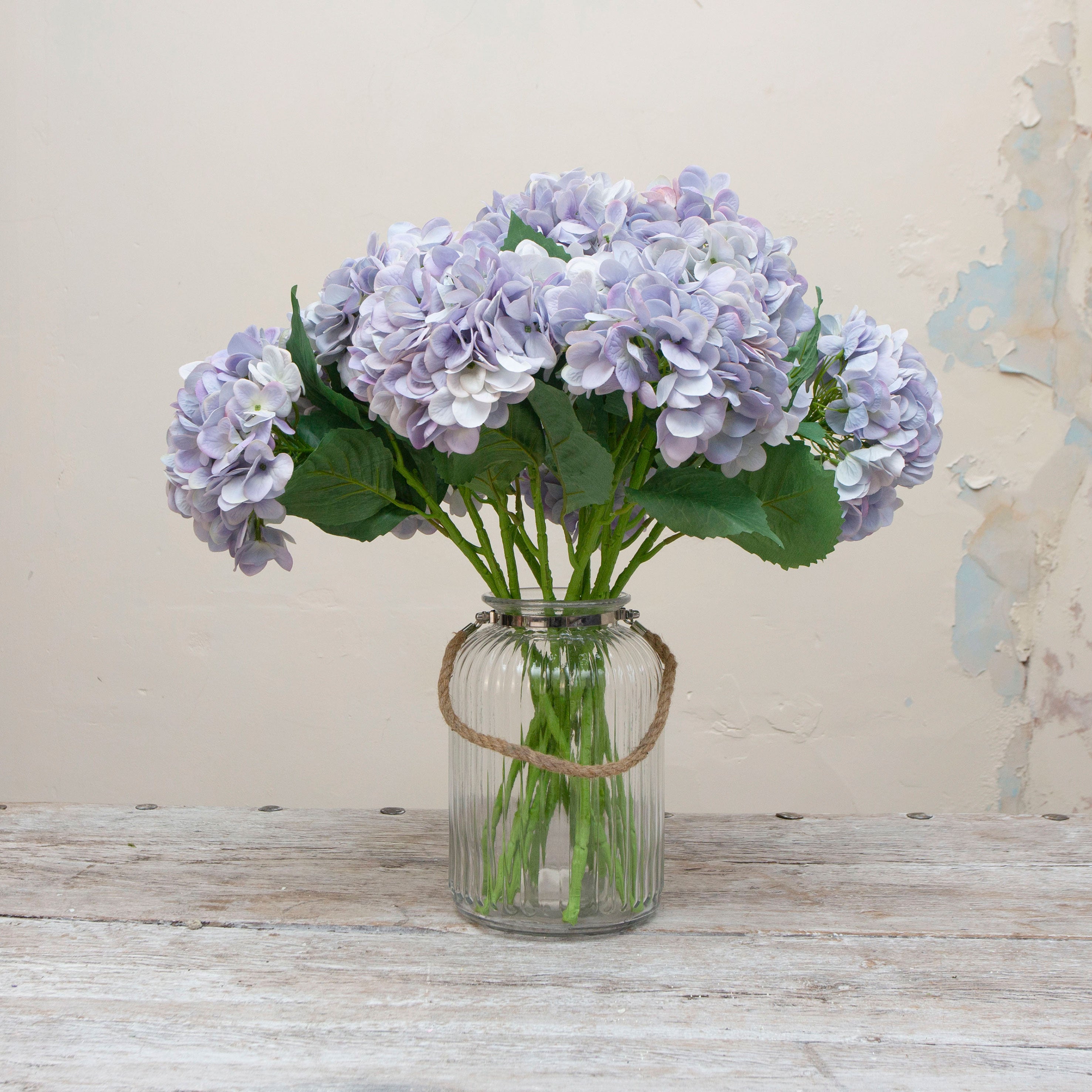 Artificial pale blue hydrangea stems displayed in a vase, showcasing a large domed flower head with delicate pastel blue tones and natural-looking green leaves