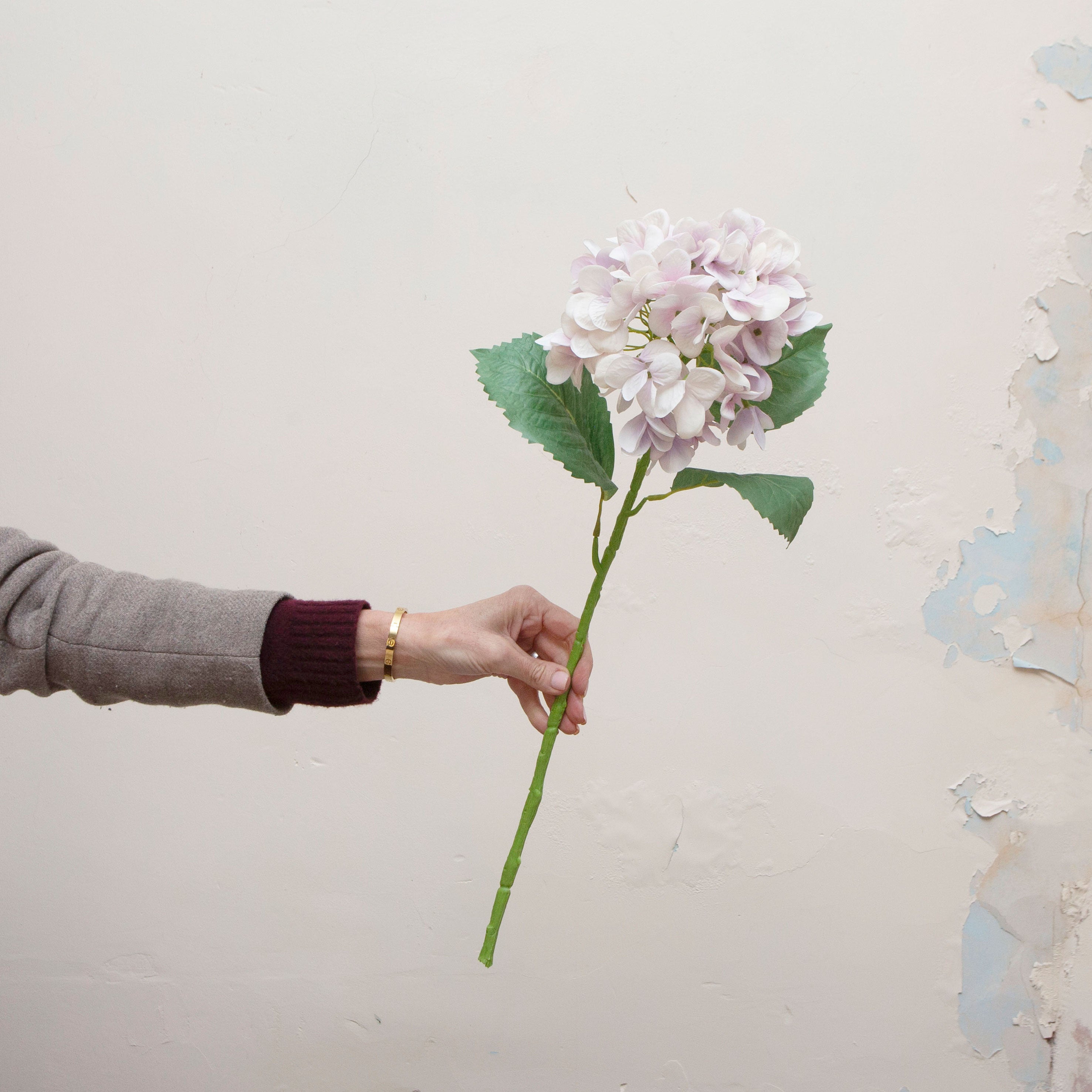 Artificial lilac hydrangea stem being held in hand, featuring a full rounded flower head with soft lilac and pale lavender petals and realistic green leaves on a sturdy stem