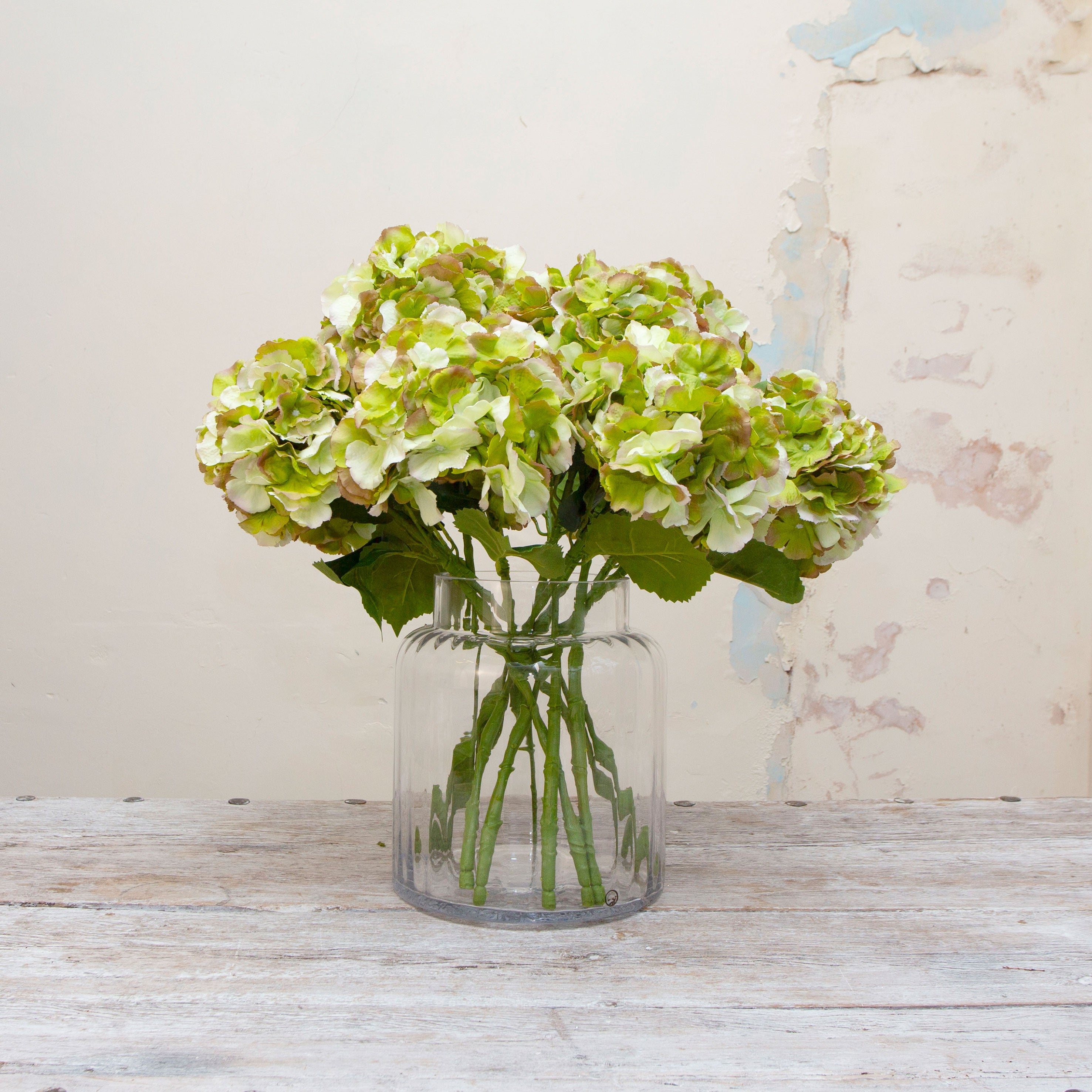 Artificial green hydrangea stem being held in hand, featuring a rounded hydrangea flower head with soft green petals and subtle blush edging, paired with realistic foliage on a sturdy stem