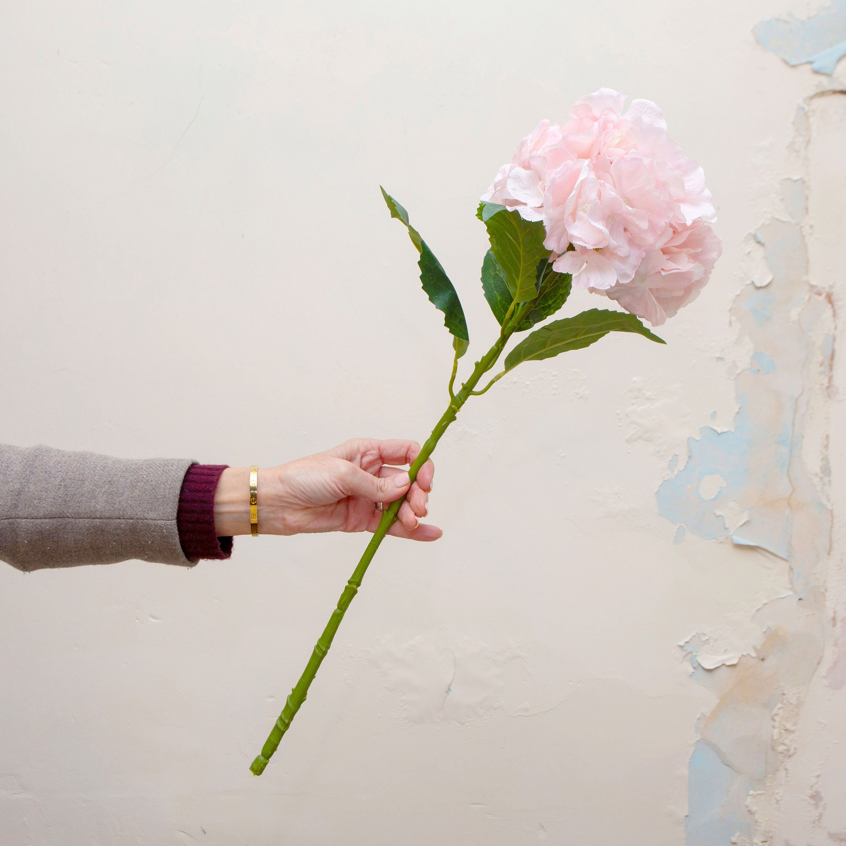 Artificial pale pink hydrangea stem being held in hand, featuring a full rounded flower head with softly layered pale pink petals and realistic green foliage on a sturdy stem.