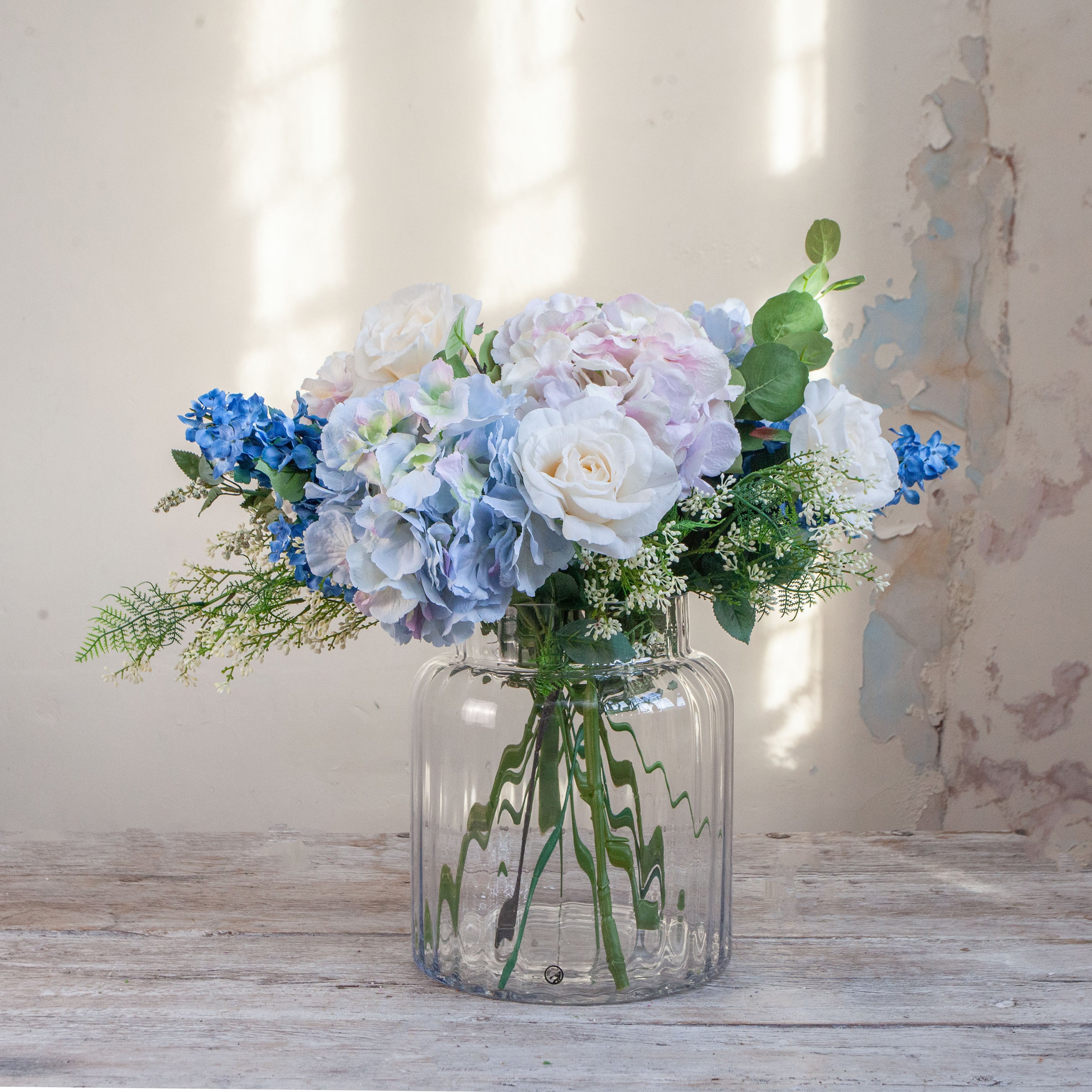 Artificial hydrangea and rose arrangement with soft greenery displayed in a large clear glass Chelsea vase,