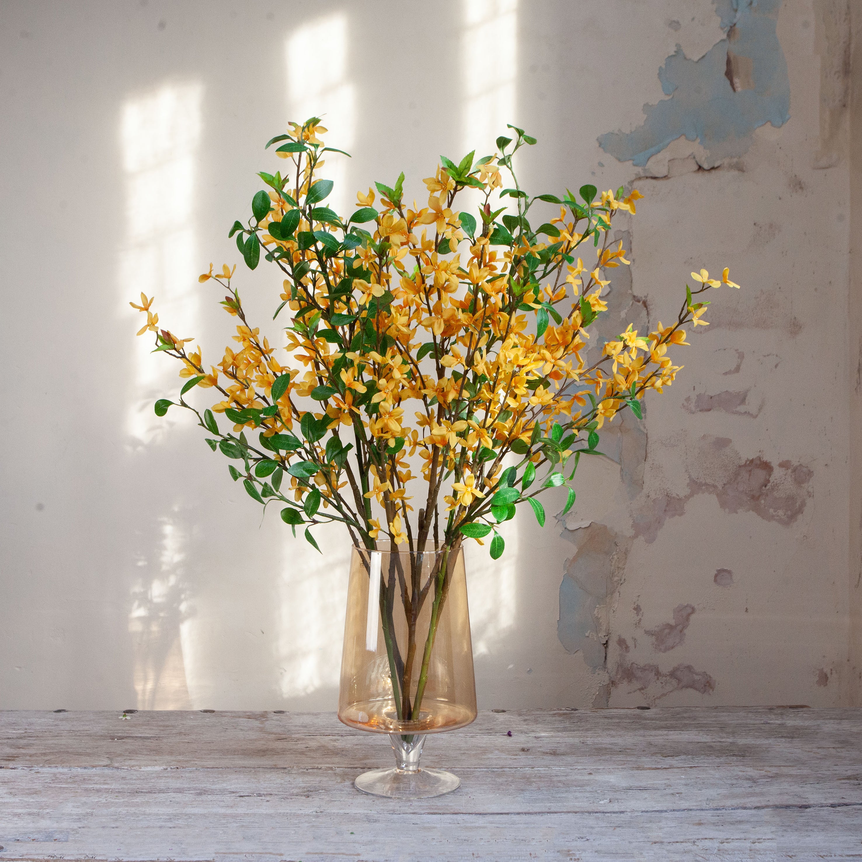 Artificial forsythia arrangement with bright yellow spring blossoms displayed in a footed amber glass vase