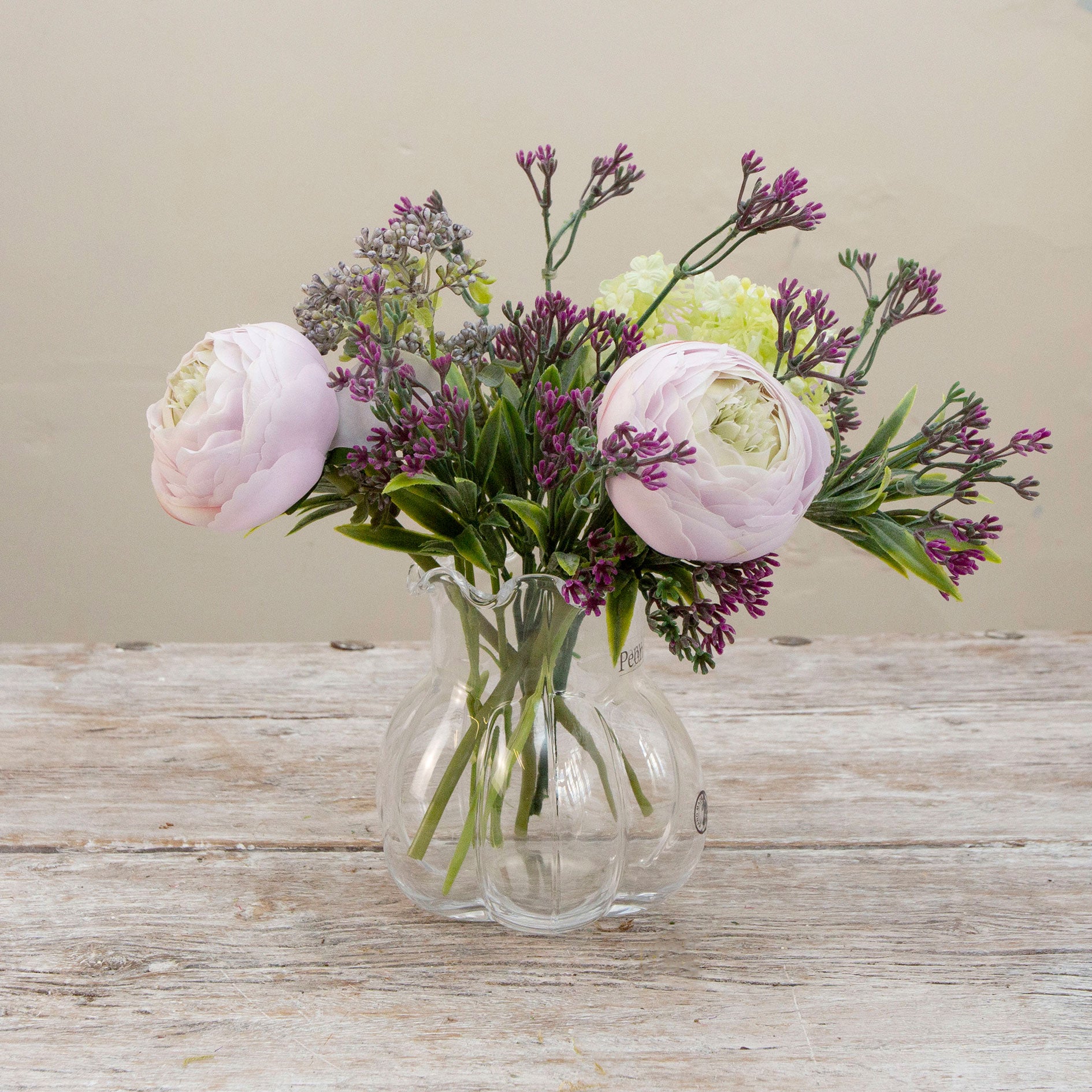 Artificial ranunculus and foliage arrangement in a clear glass garlic vase