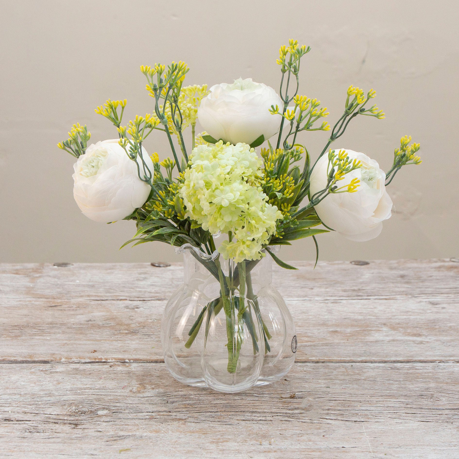 Artificial ranunculus and foliage arrangement in a clear glass garlic vase
