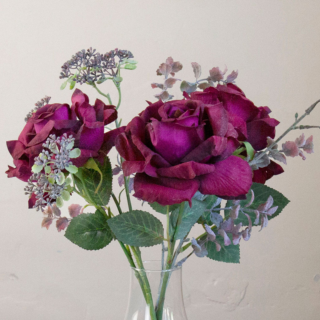 Close-up of artificial burgundy roses and patrinia in a clear glass milk bottle vase.