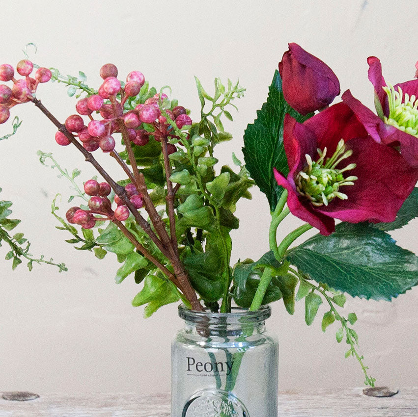Close-up of an artificial burgundy hellebore and berry arrangement with green foliage in a glass bottle vase.