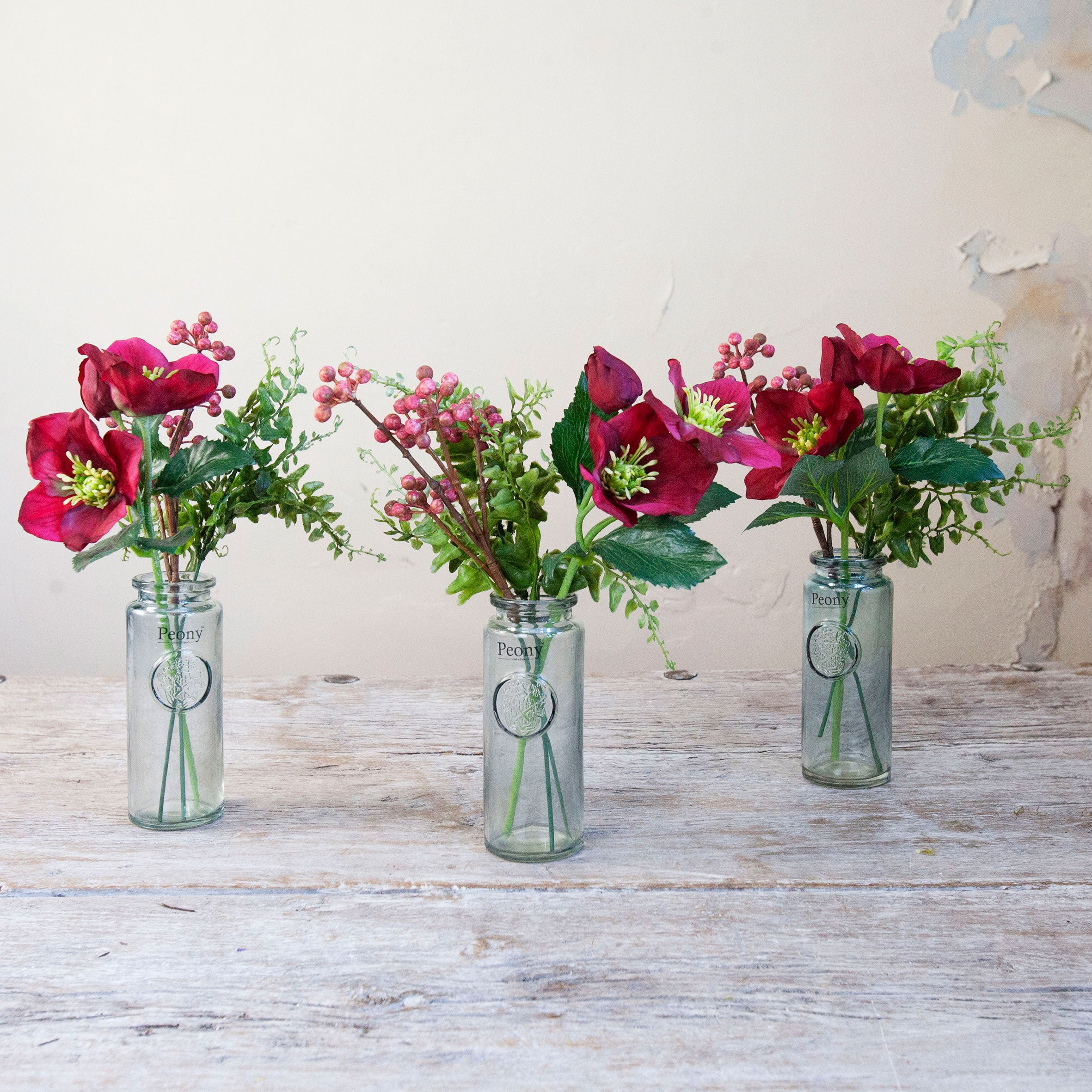 Set of three artificial burgundy hellebore and berry arrangements with green foliage in individual glass bottle vases.
