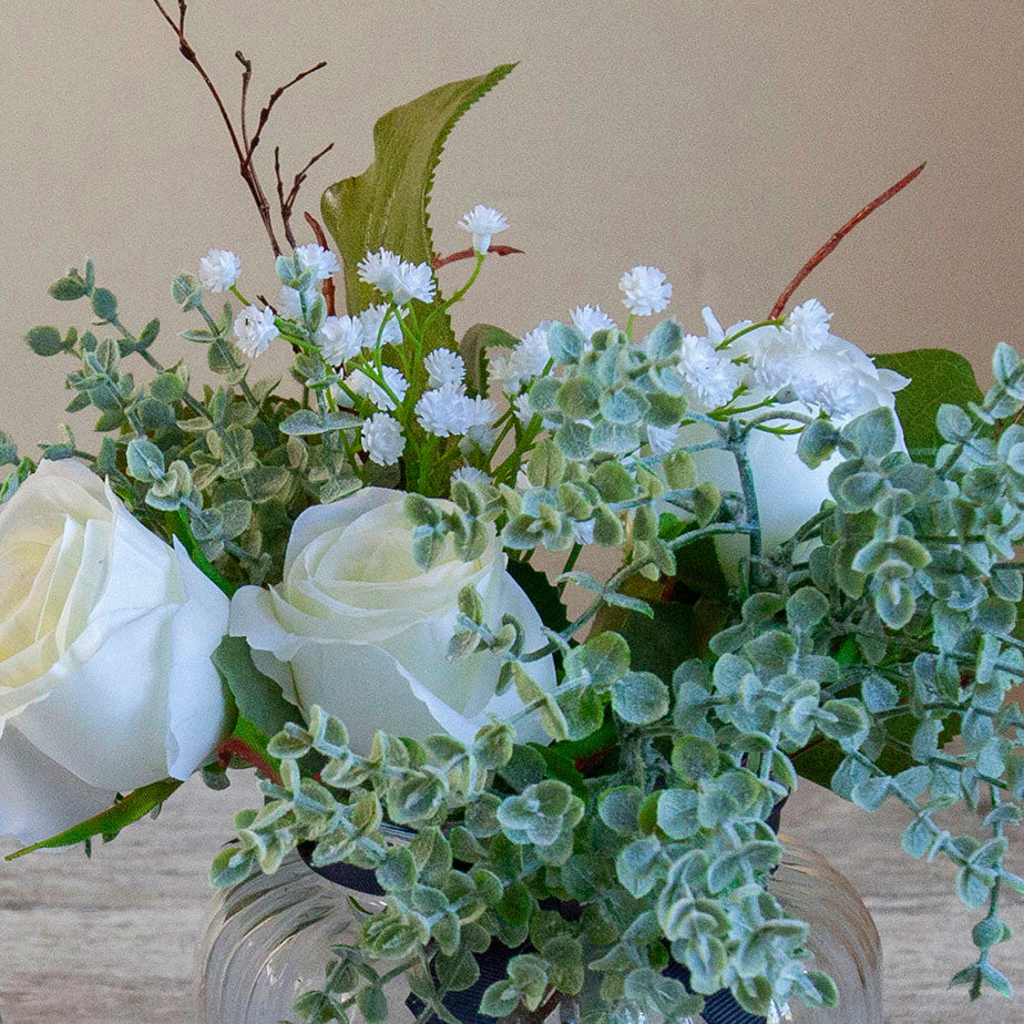 Close-up of artificial white roses, mini eucalyptus and gypsophila set in a clear optic glass jar vase.
