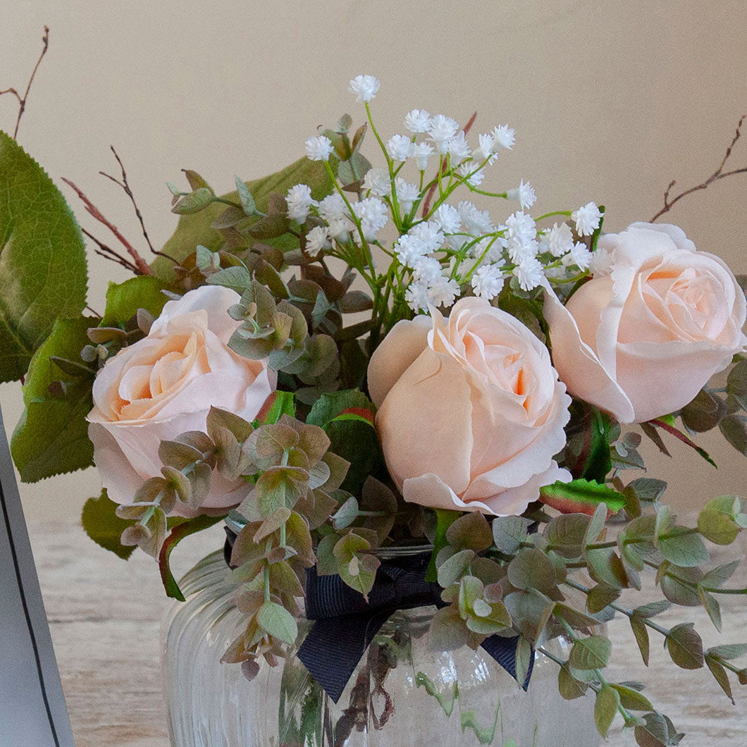 Close-up of artificial blush roses, mini eucalyptus and gypsophila set in a clear optic glass jar vase.