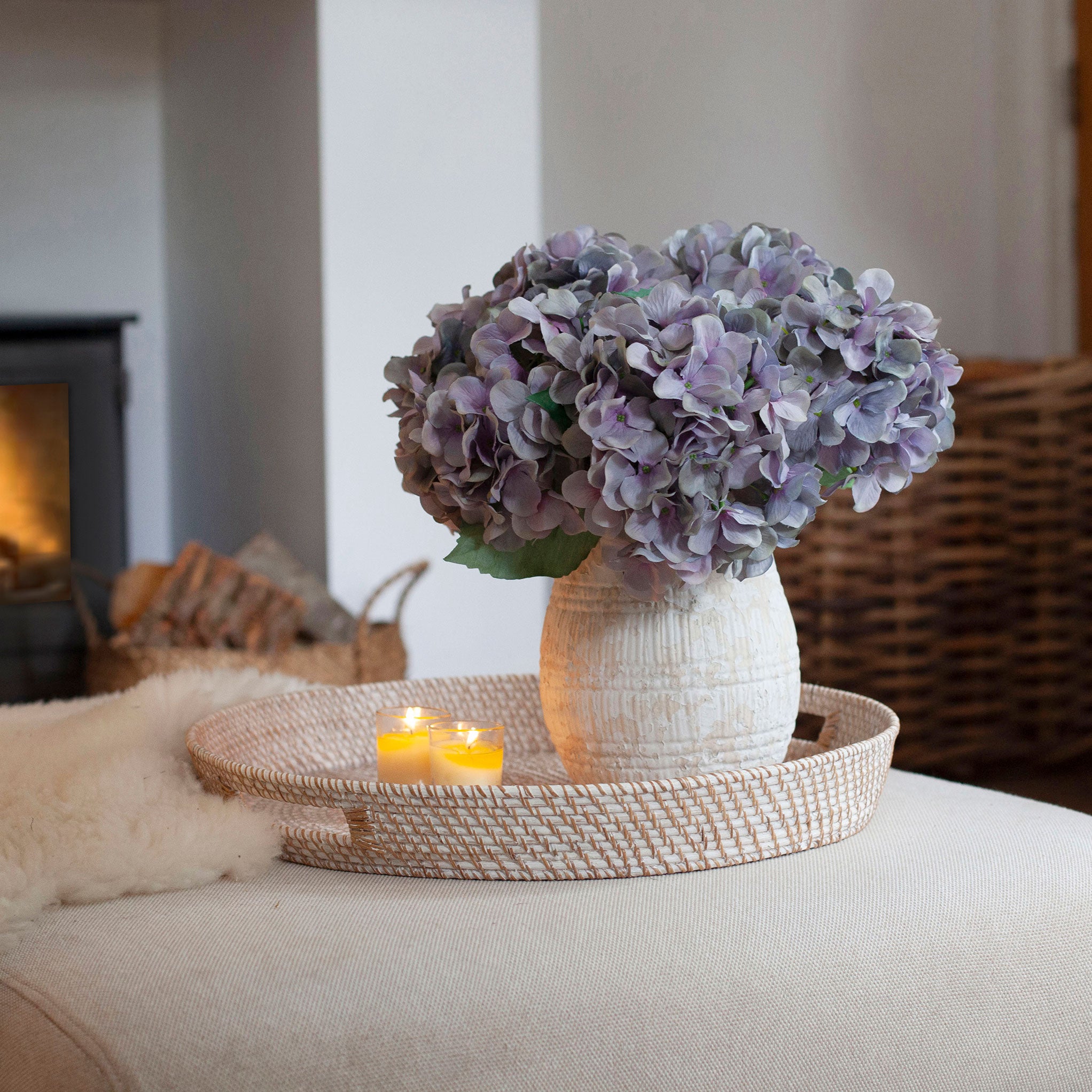 Artificial hydrangea arrangement in soft blue, lilac and grey tones in a rustic textured white pot, styled on a woven tray in a cosy living room.