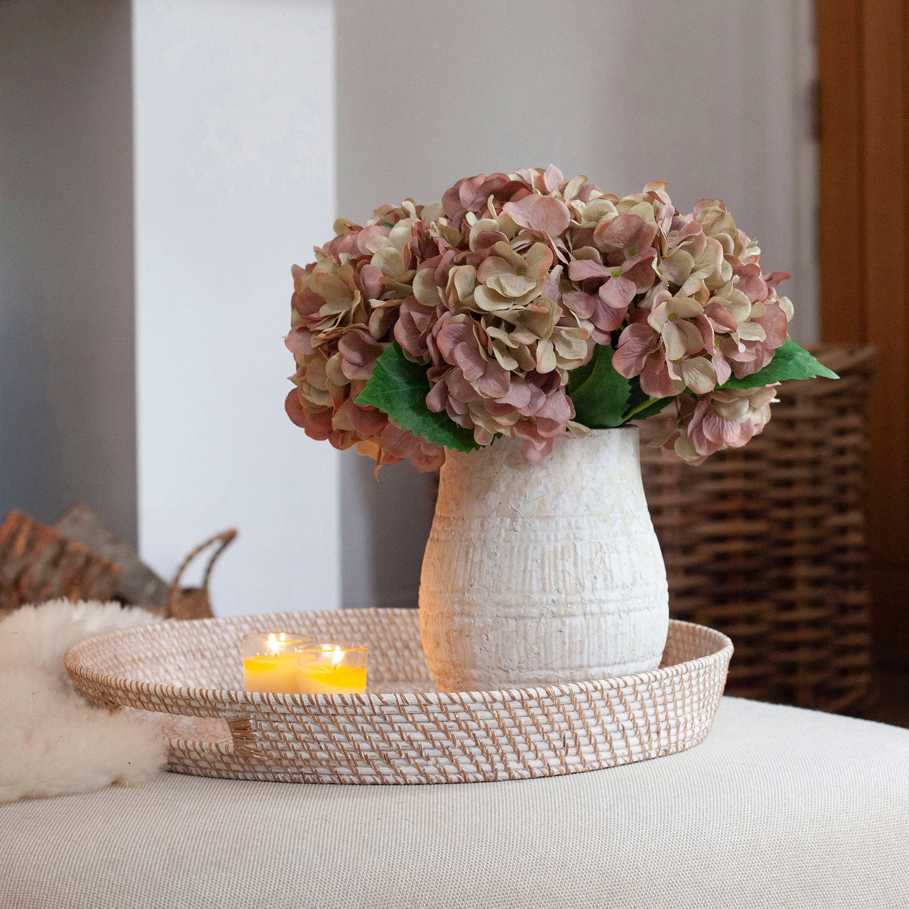 Artificial hydrangea arrangement in soft blush, mauve and green tones in a rustic textured white pot, styled on a woven tray in a cosy living room.