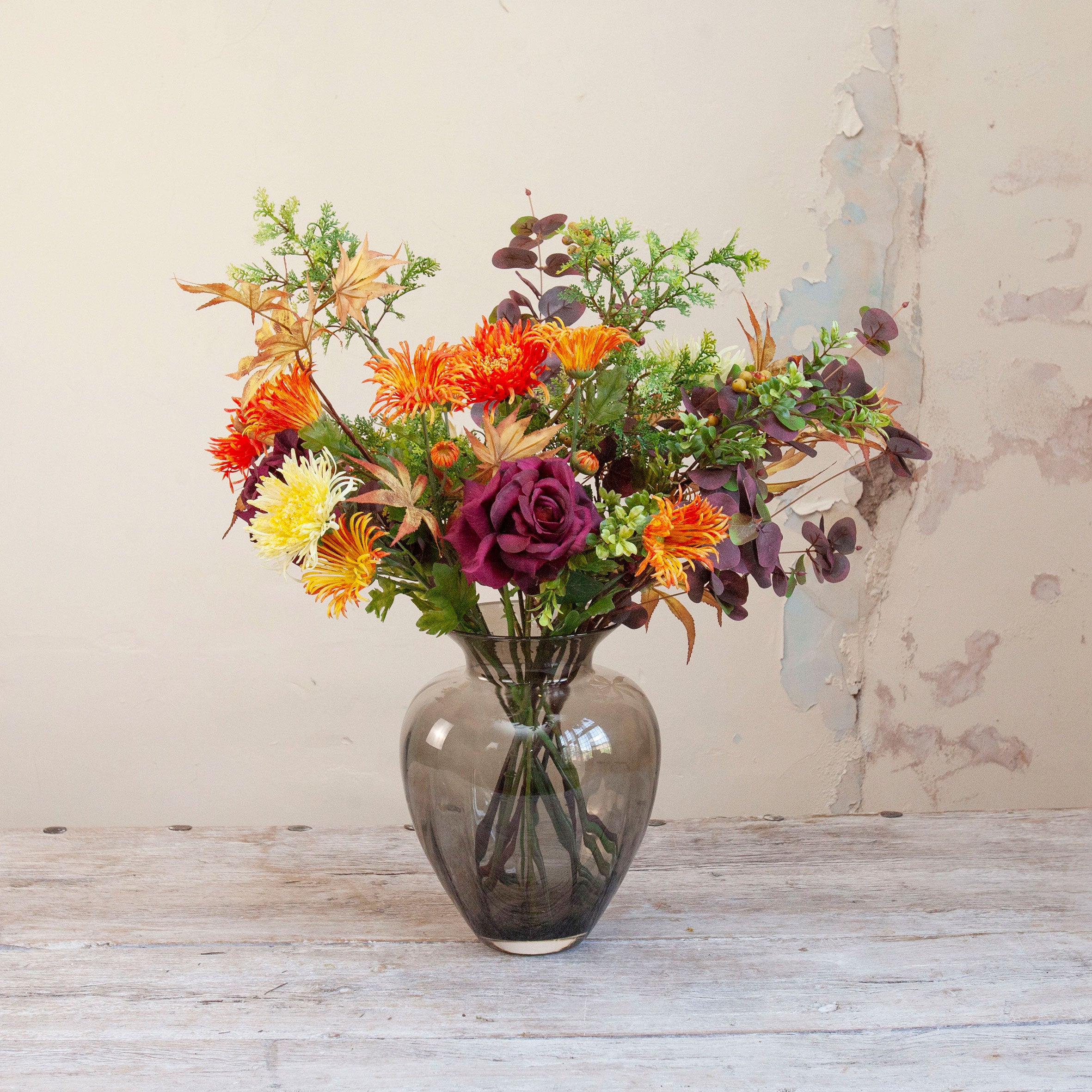 Artificial chrysanthemum and autumn foliage bouquet in rich orange, burgundy and green tones, displayed in a smoked glass vase.