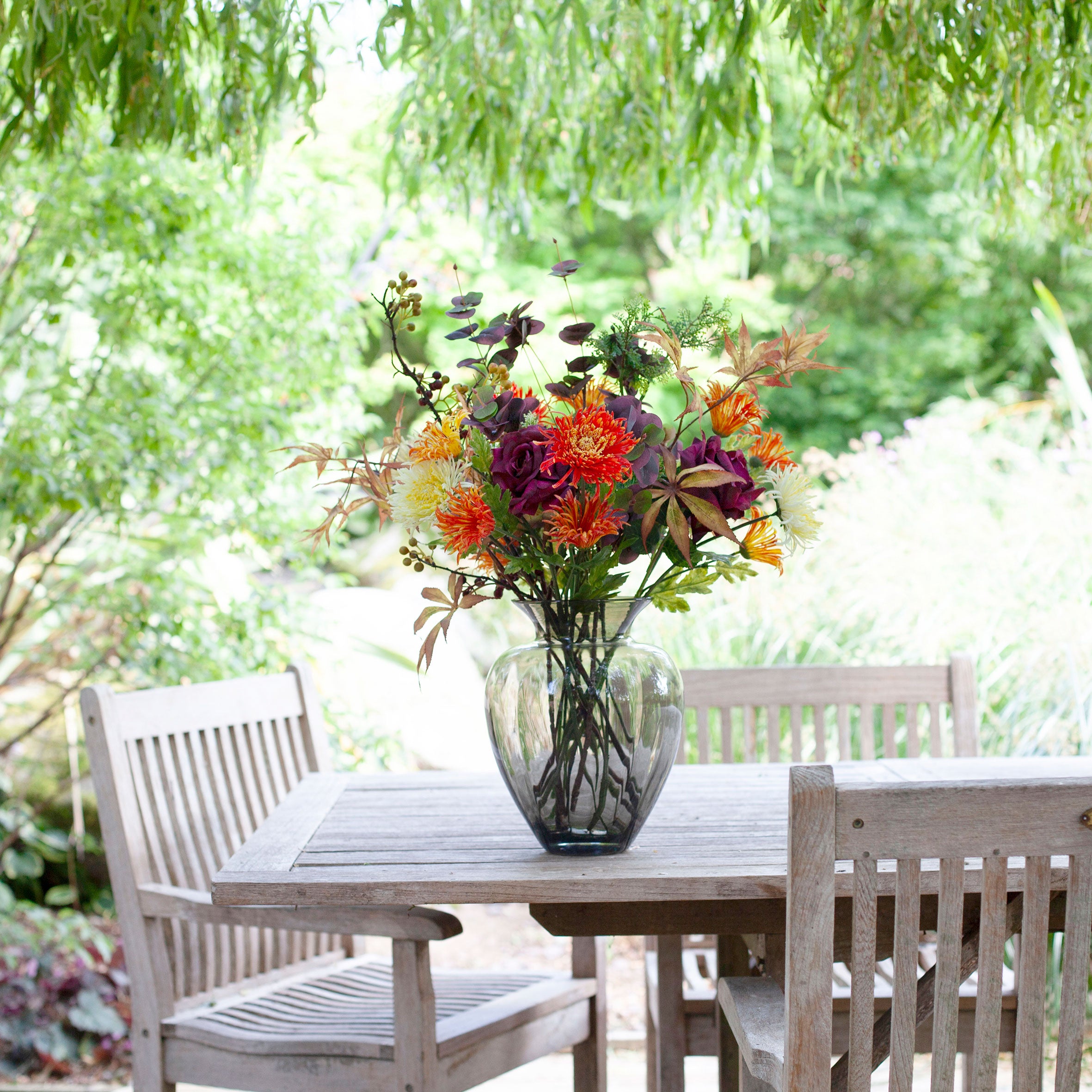 Artificial chrysanthemum and autumn foliage bouquet in rich orange, burgundy and green tones, displayed in a smoked glass vase on an outdoor garden table.