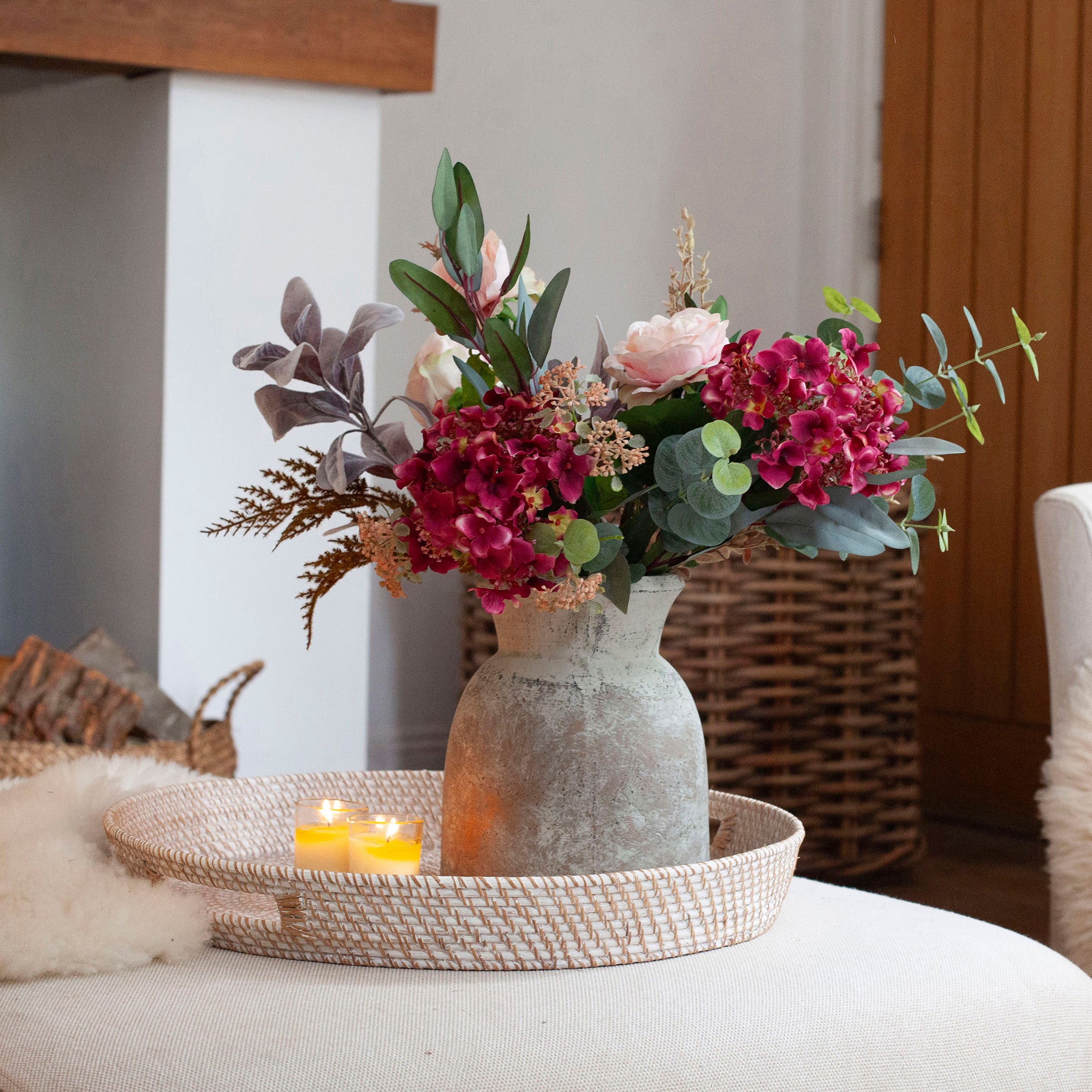 Artificial rose and hydrangea arrangement in soft pink and rich burgundy tones with eucalyptus and autumn foliage, styled in a rustic vase in a cosy living room.
