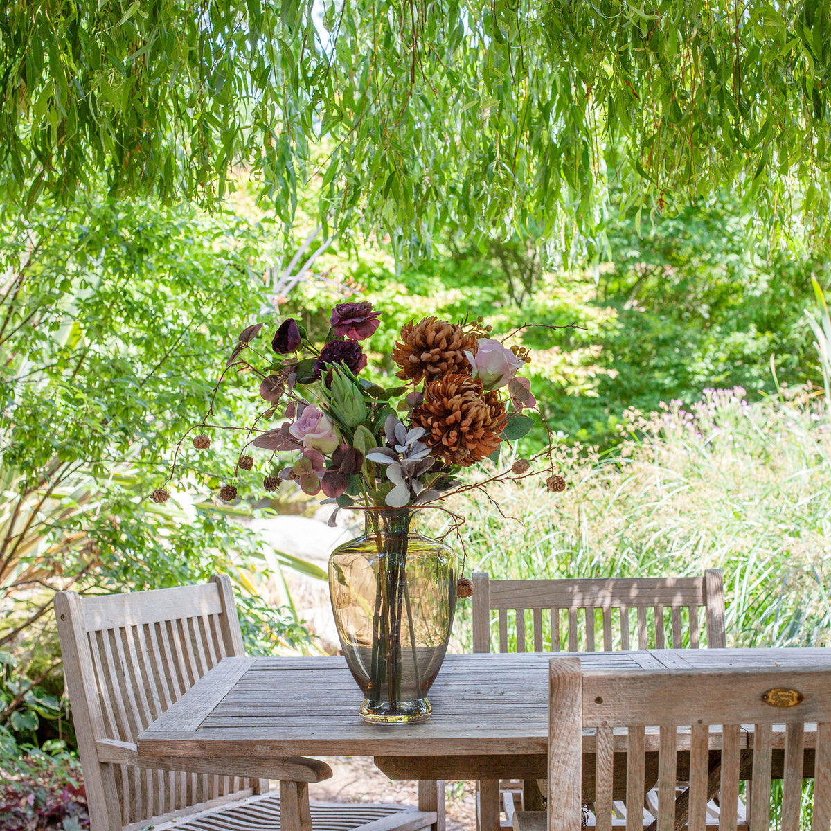 Artificial chrysanthemum, rose and foliage bouquet in warm autumn tones, displayed in an amber glass vase on an outdoor garden table.