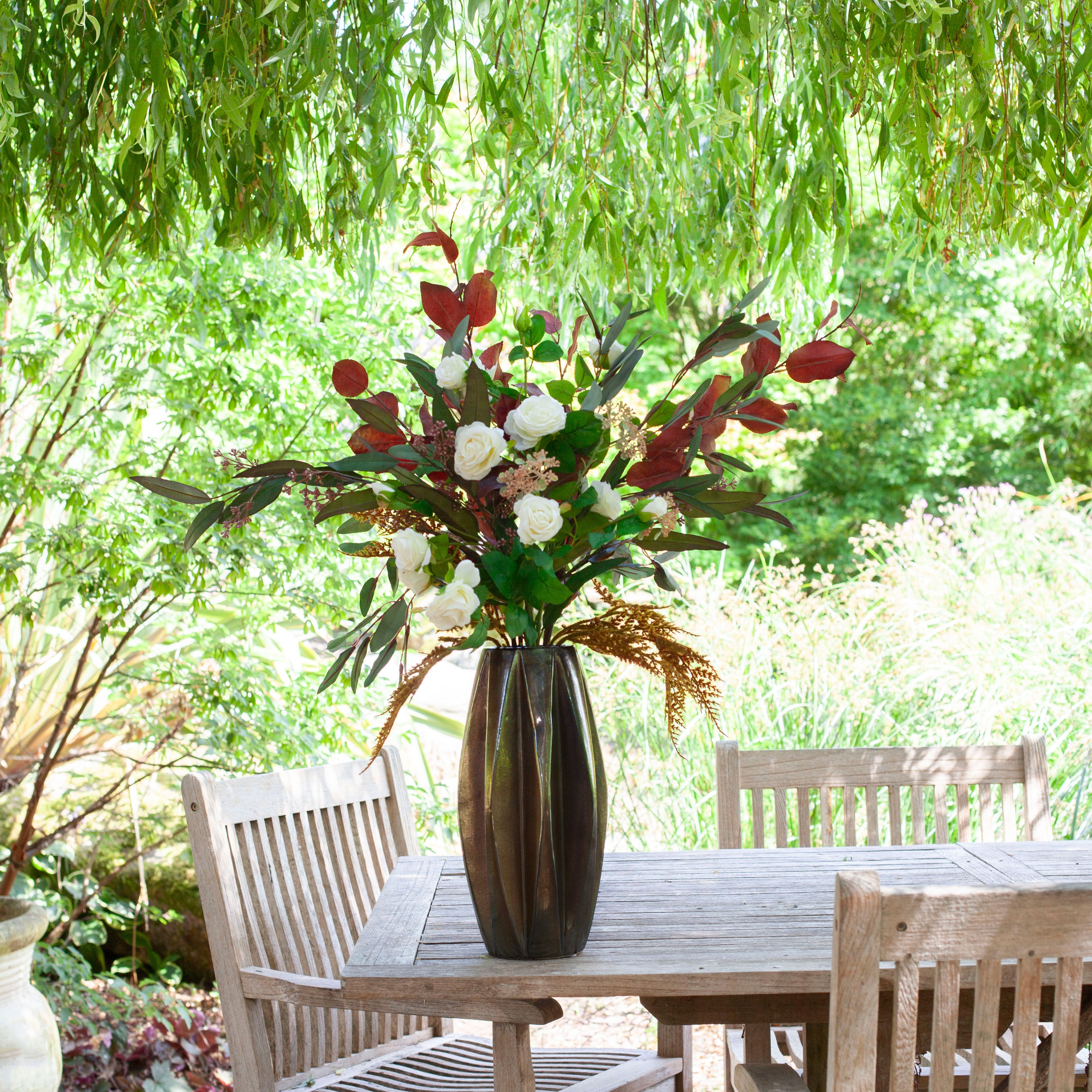 Artificial rose and berry arrangement with white roses and rich autumn foliage in a tall bronze geometric pot, styled on an outdoor garden table.