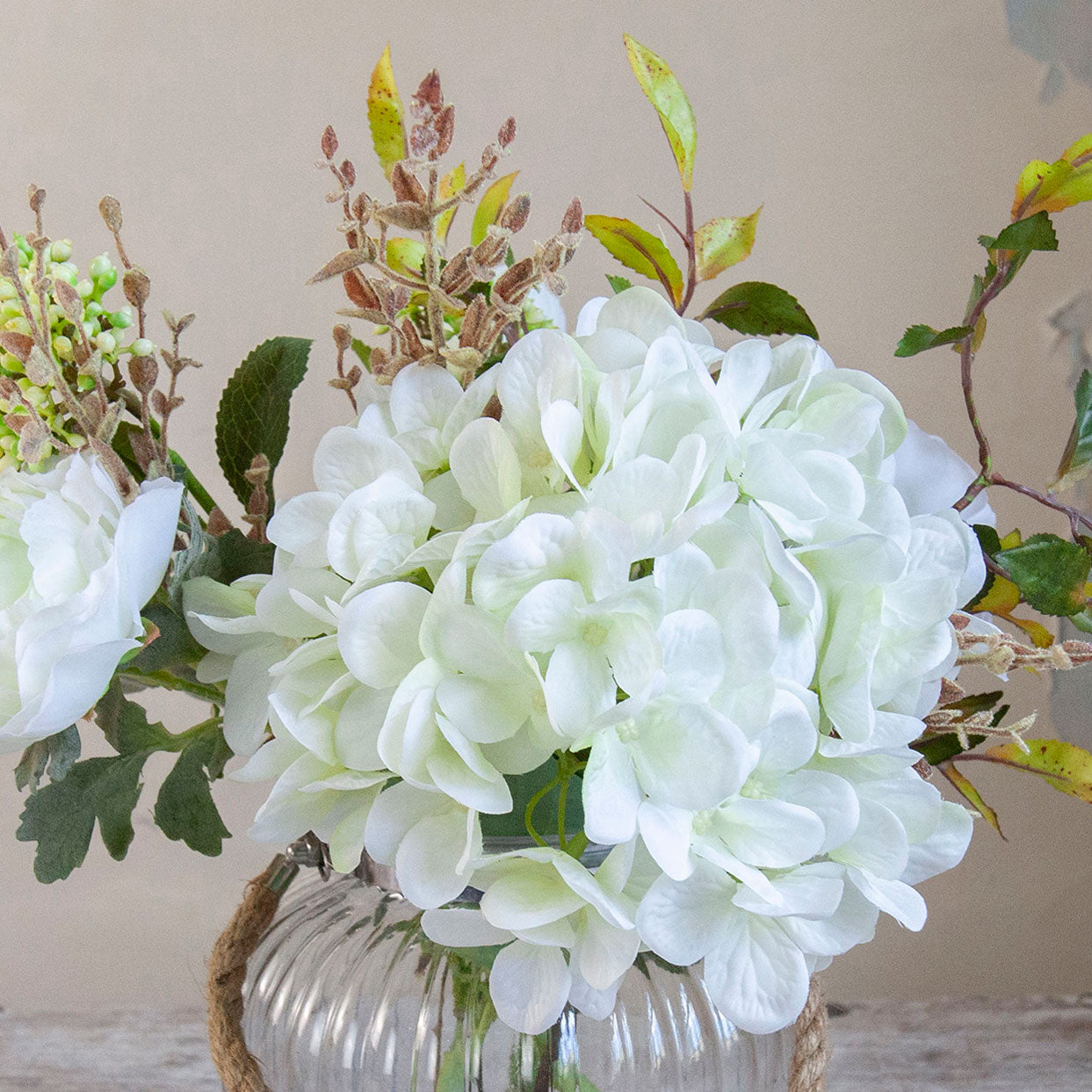 Close-up of artificial white hydrangea and ranunculus flowers with realistic autumn foliage in a clear glass rope lantern vase
