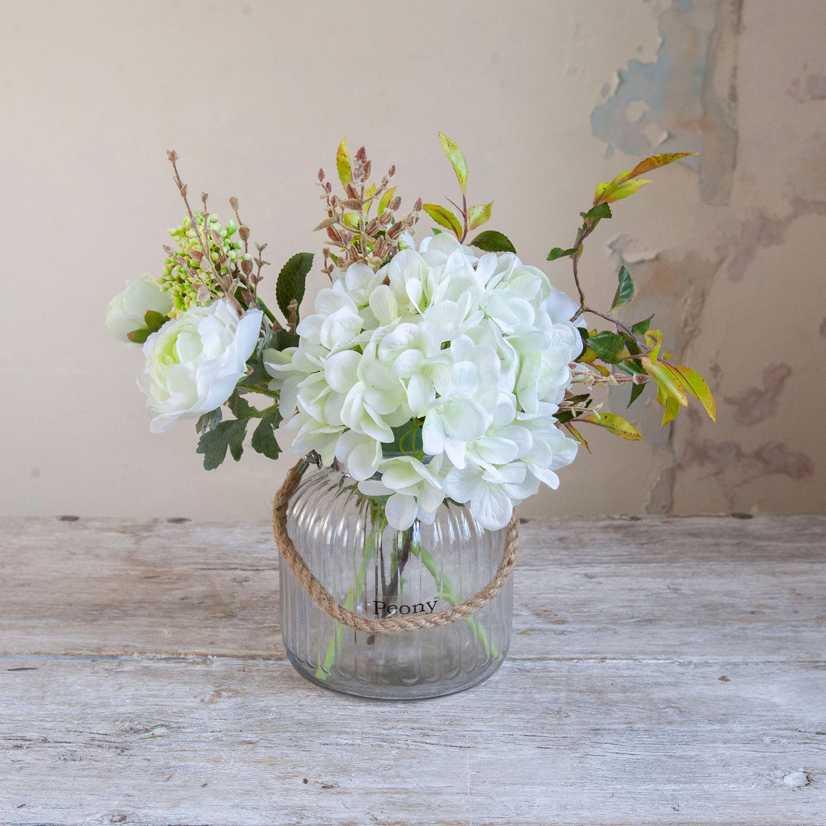Artificial hydrangea, ranunculus and foliage arrangement in soft white and green tones, arranged in a medium clear glass rope lantern vase.