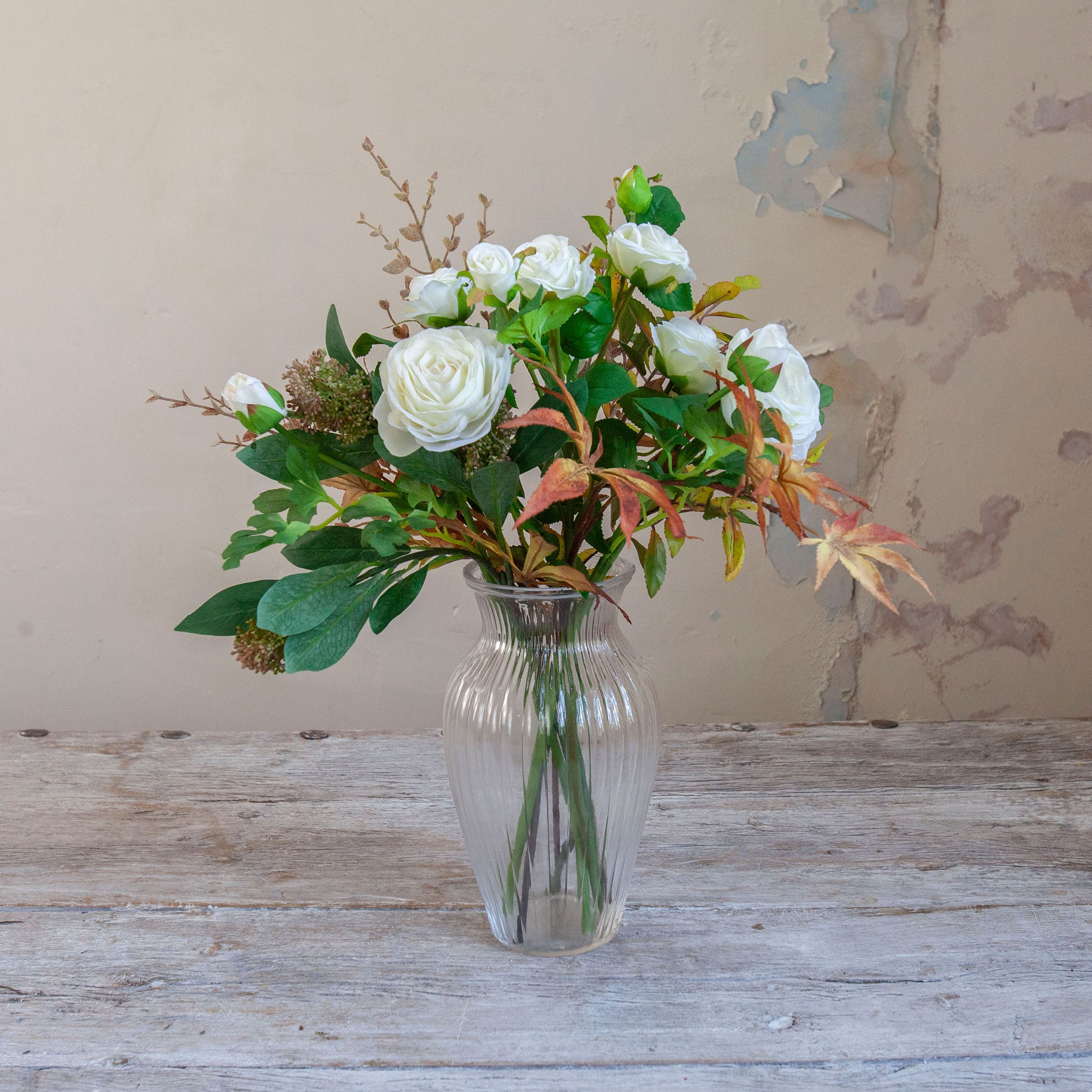Artificial rose, ranunculus and autumn foliage bouquet with white flowers and warm seasonal leaves, displayed in a clear glass vase.