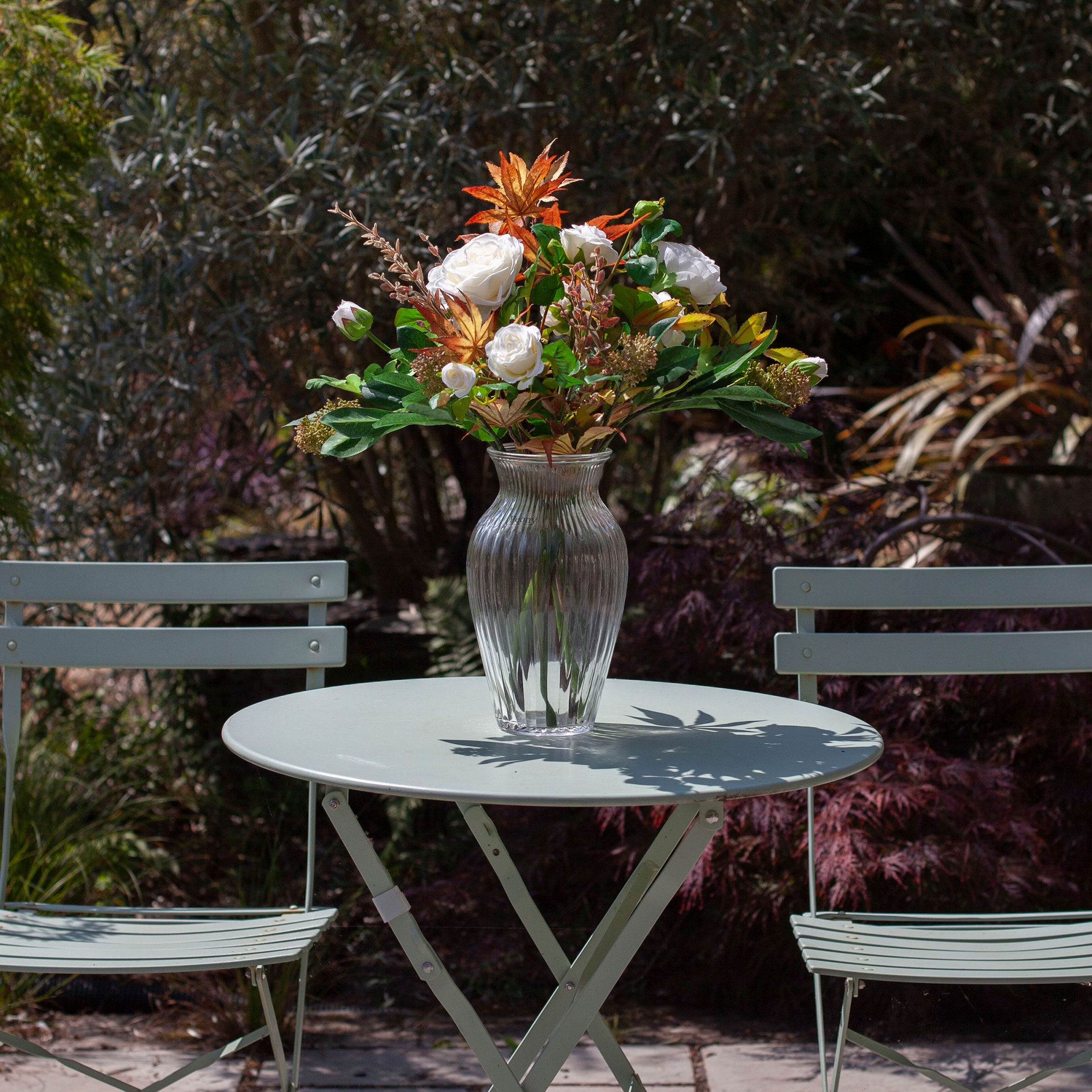 Artificial rose, ranunculus and autumn foliage bouquet with white flowers and warm seasonal leaves, displayed in a clear glass vase on an outdoor garden table.