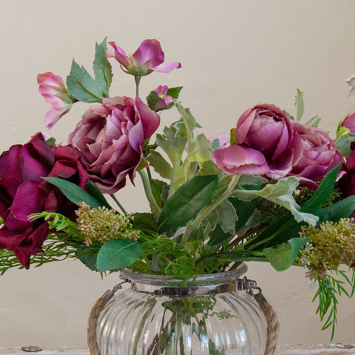 Close-up of artificial roses, hellebores and foliage in rich burgundy and mauve tones, displayed in a clear glass rope lantern vase.