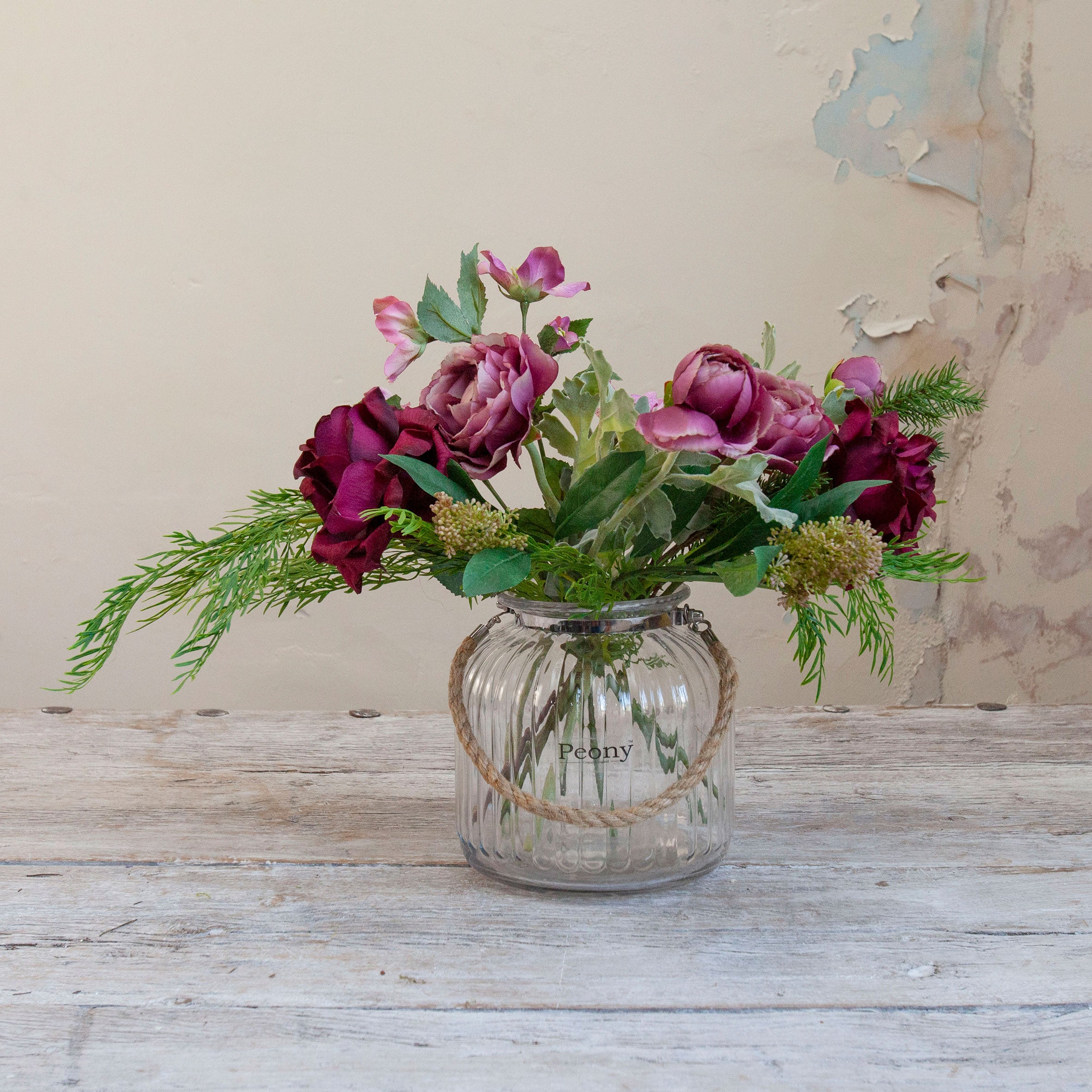 Artificial roses, hellebores and foliage bouquet in rich burgundy and mauve tones, displayed in a clear glass rope vase.