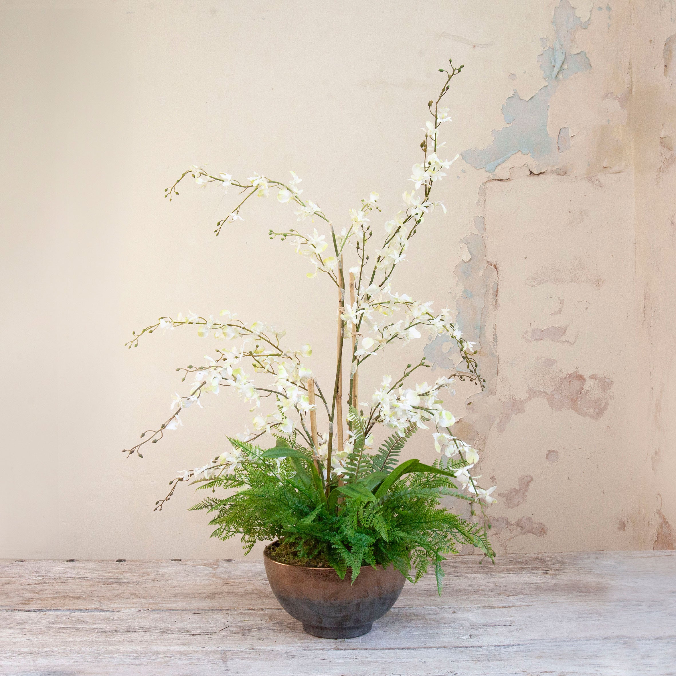 Artificial white dancing orchids with fern foliage arranged in a bronze footed bowl.