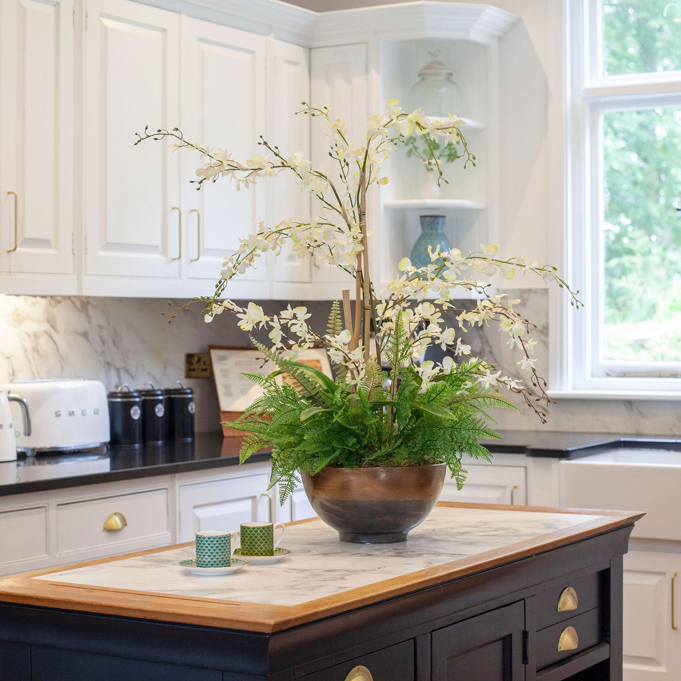 Artificial white dancing orchids with fern foliage arranged in a bronze footed bowl, styled on a kitchen island.