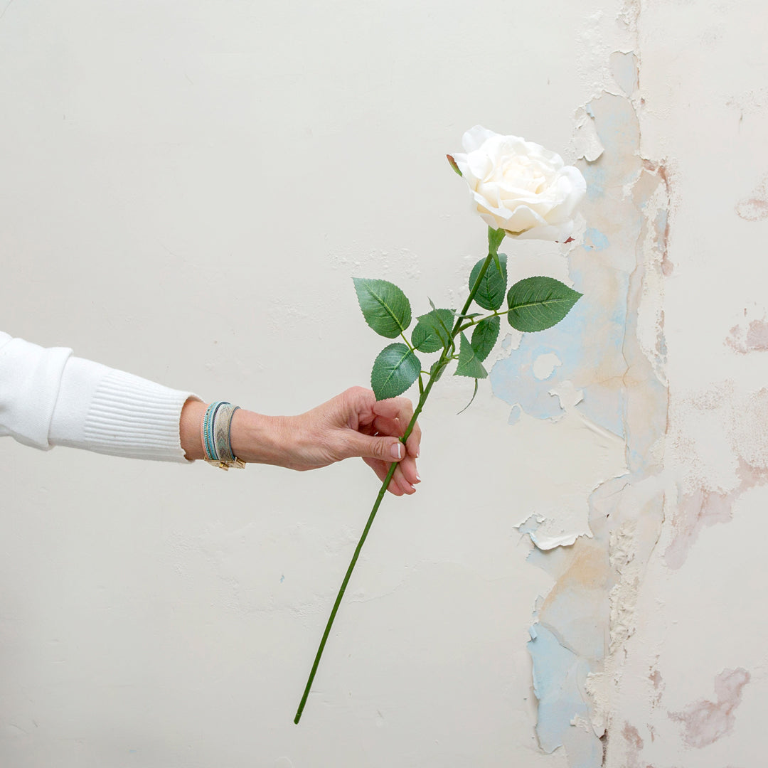 Artificial white rose stem being held in hand, featuring a single open rose bloom in soft bridal white with realistic green leaves on a long slender stem