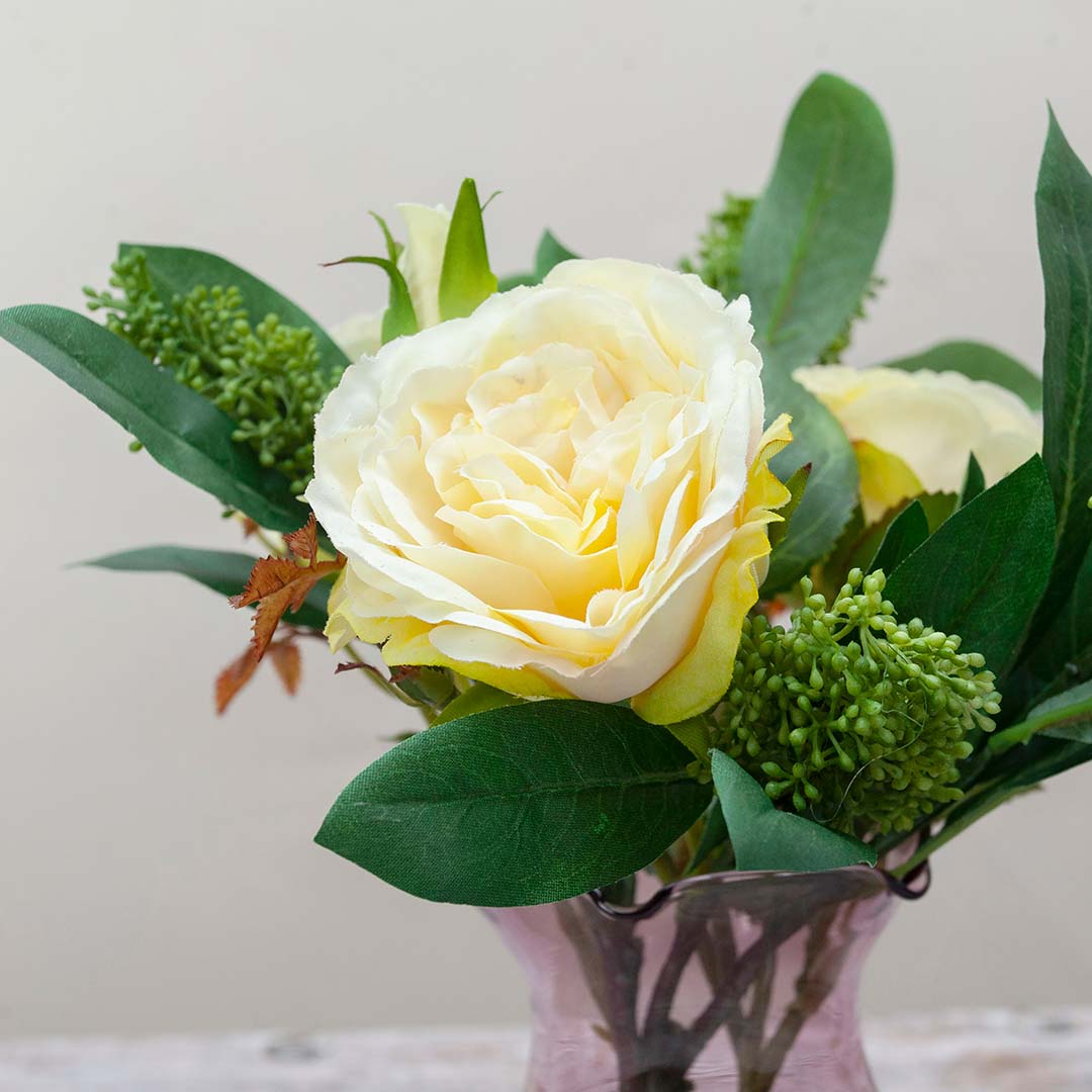 Artificial rose and skimmia arrangement displayed in a modern Prisma vase, presented with a gift bag and a 25ml rose fragrance bottle.