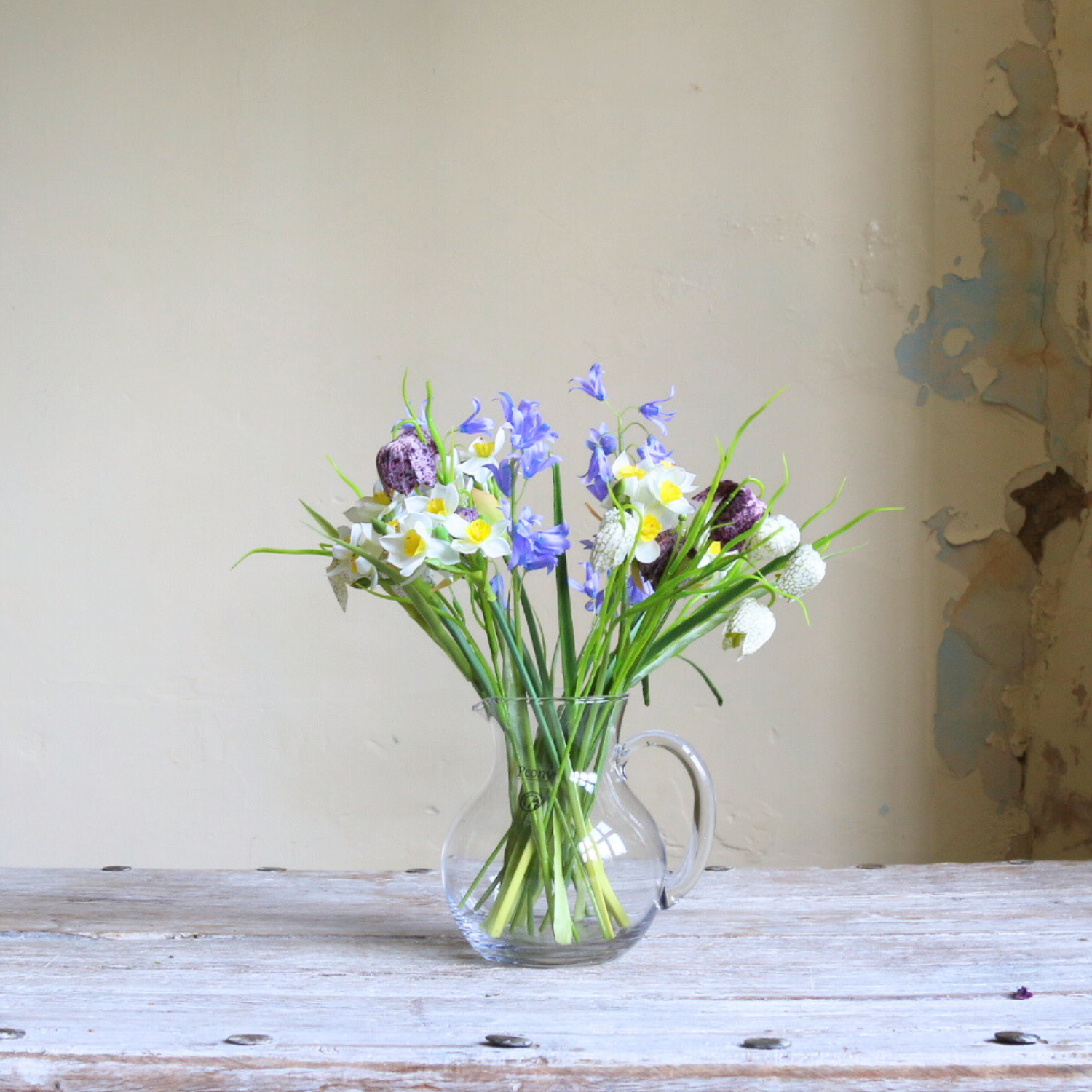 Artificial spring flower arrangement with daffodils, bluebells, and fritillaria arranged in a clear glass jug.