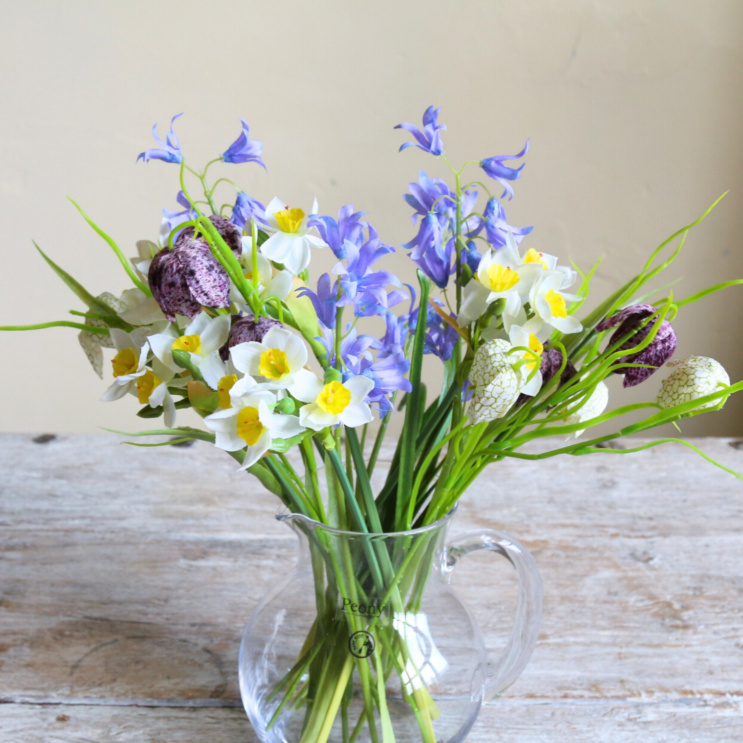 Artificial spring flower arrangement with daffodils, bluebells, and fritillaria arranged in a clear glass jug.