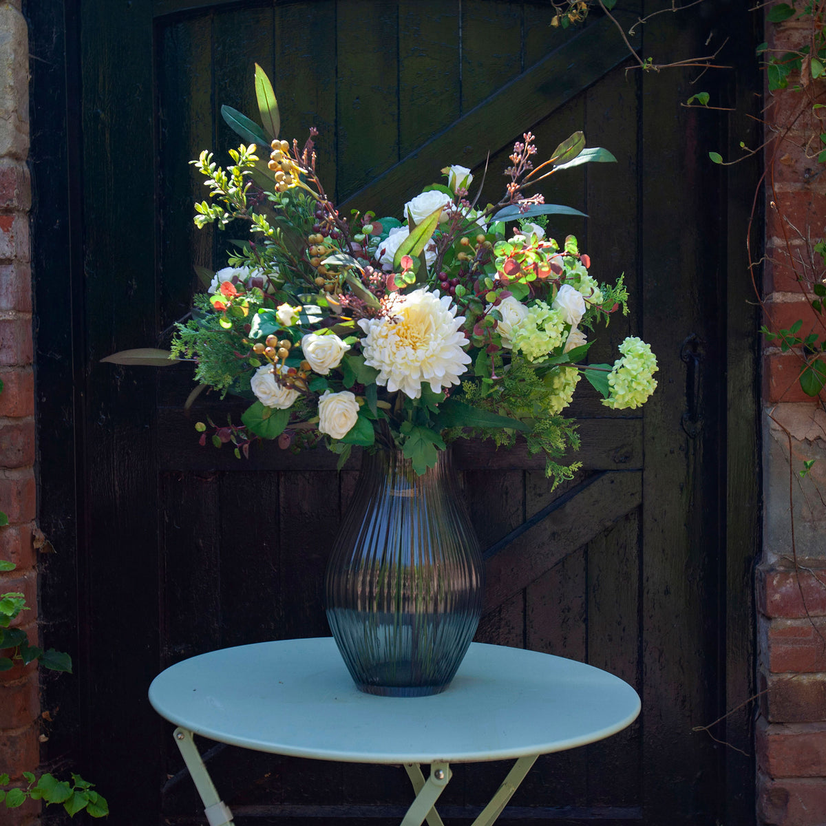 Artificial cream chrysanthemums, roses and green viburnum with berries and botanical foliage, arranged in a tall smoked optic glass vase against a rustic dark wooden backdrop.