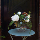 Artificial white chrysanthemums and cream roses with autumn foliage, fern leaves and dark berries, arranged in a clear round glass bowl.