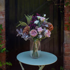 Artificial garden roses, burgundy orchids and bronze chrysanthemums with silvery foliage, arranged in a tall clear glass vase against a rustic dark wooden backdrop.