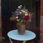 Artificial burgundy peony, soft peach spray roses and purple thistles with rich autumn foliage, arranged in a tall rustic textured vase against a dark wooden backdrop.