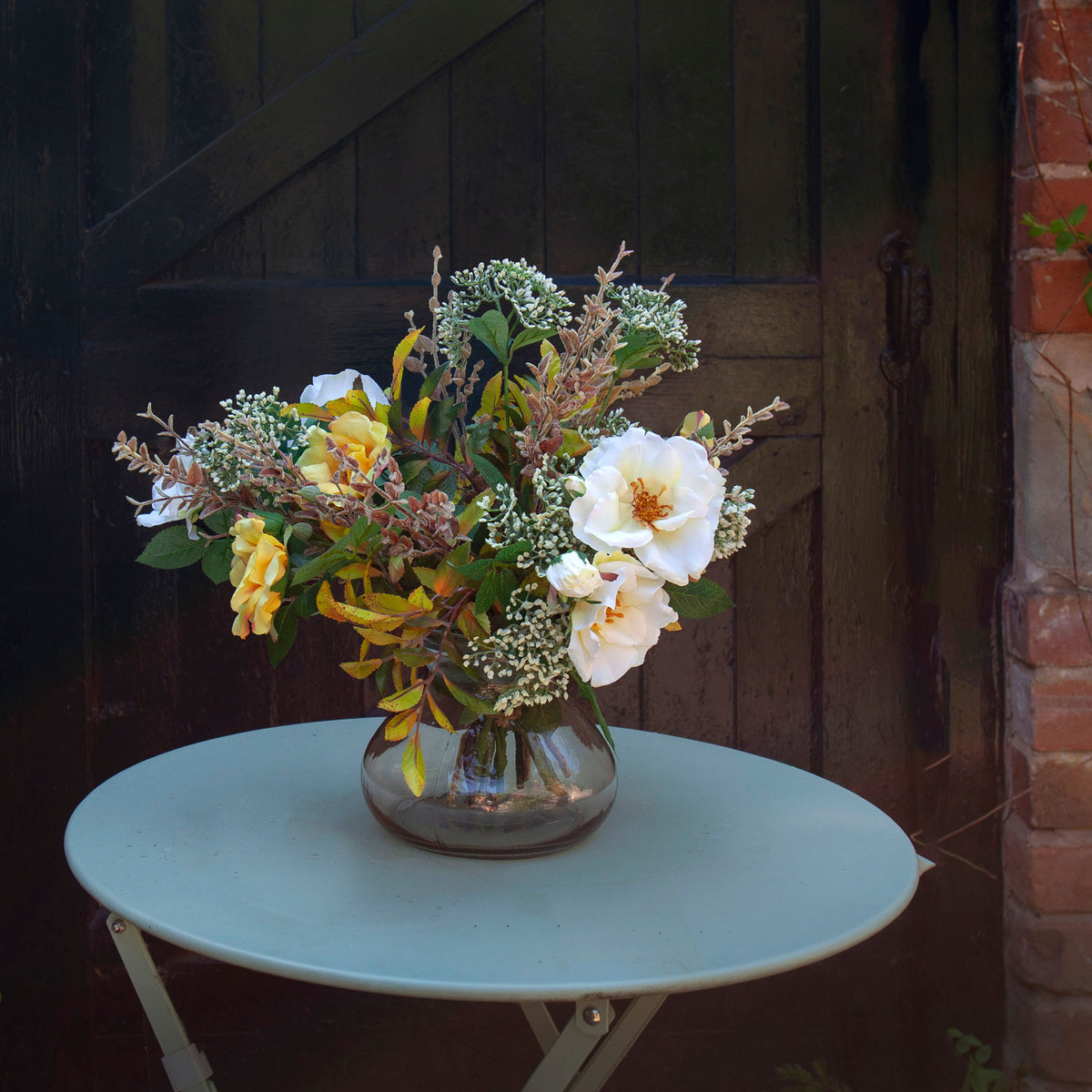 Artificial yellow and ivory roses, skimmia and seasonal foliage arrangement displayed in a smoked optic glass vase on a rustic wooden table against a warm neutral background.