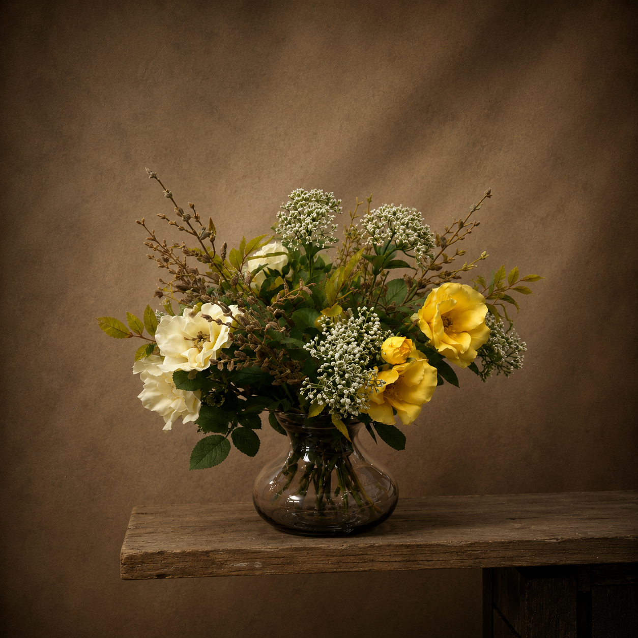 Artificial yellow and ivory roses, skimmia and seasonal foliage arrangement displayed in a smoked optic glass vase on a rustic wooden table against a warm neutral background.