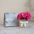 Artificial pink roses in a pearl-patterned decorative pot with cream bow, displayed beside a silver Peony gift bag.