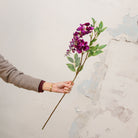 Artificial purple wisteria stem being held in hand, featuring cascading clusters of soft purple blooms with delicate petal detail on a natural-looking stem