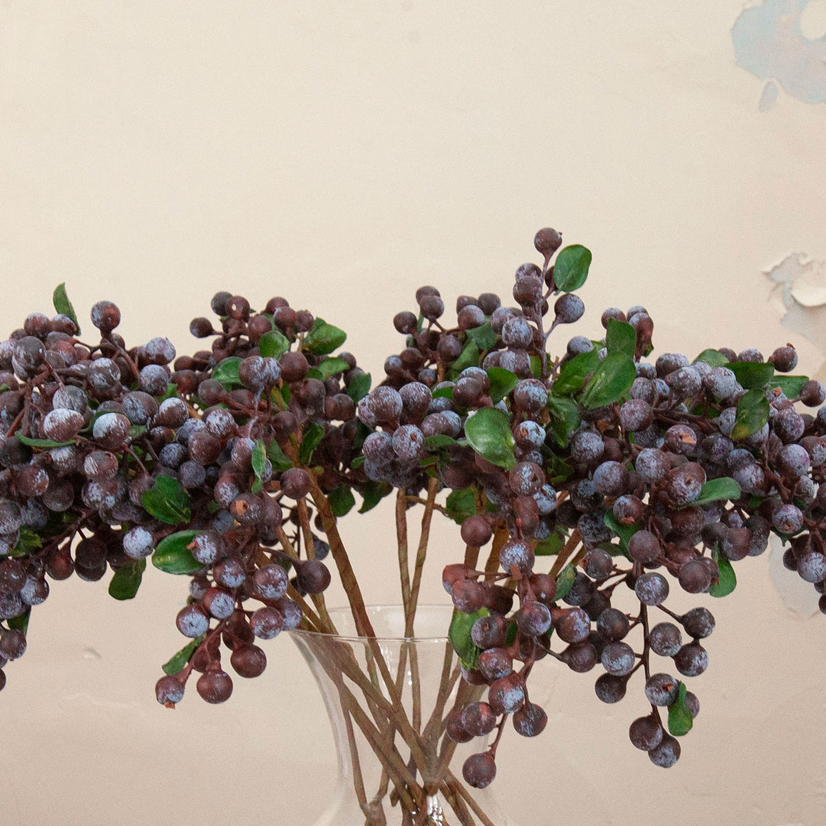 Close up of artificial purple berry cluster stems with green foliage displayed in a clear glass vase