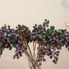 Close up of artificial purple berry cluster stems with green foliage displayed in a clear glass vase