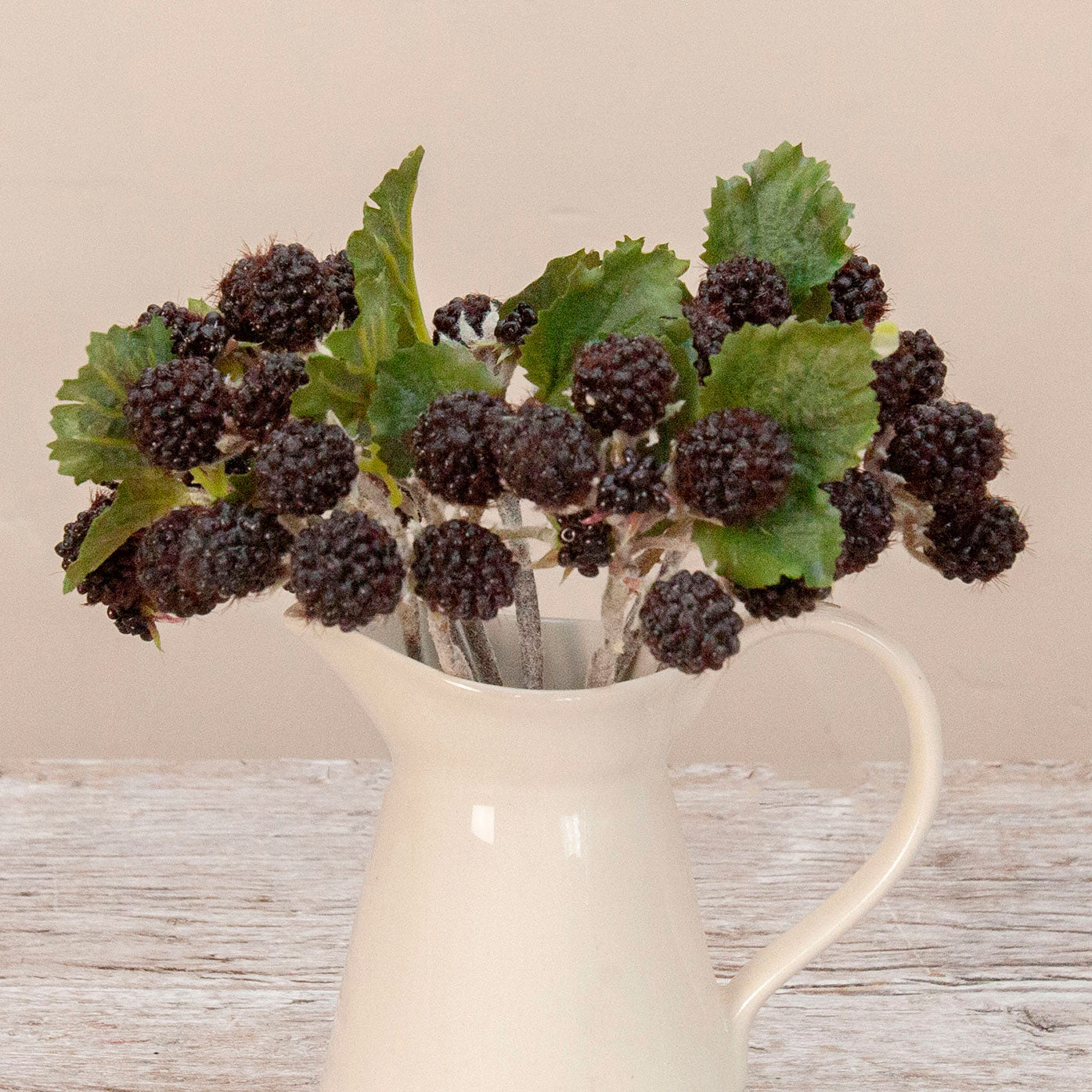 Close up of artificial blackberry cluster stems with dark berries and green leaves displayed in a cream ceramic jug