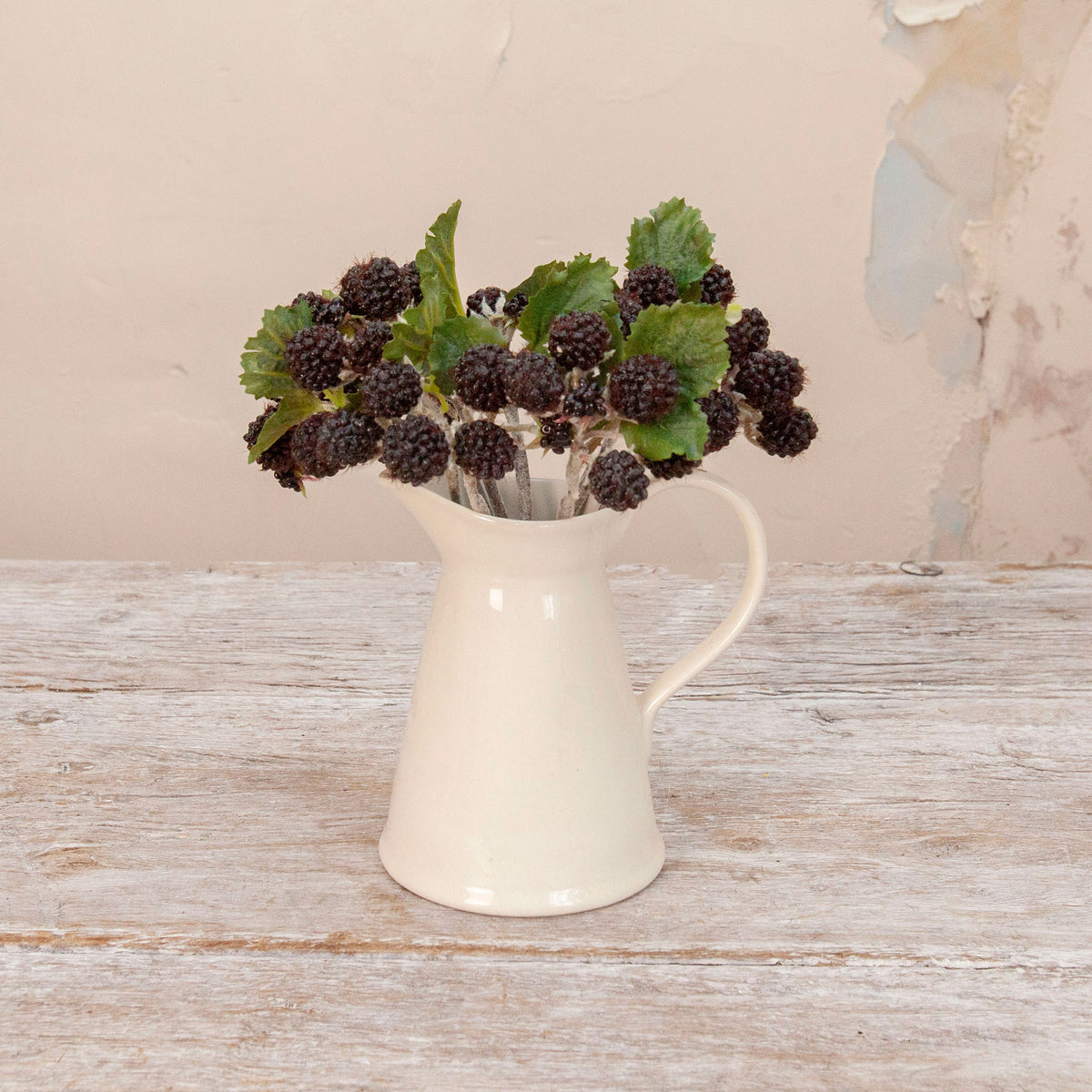 Artificial blackberry cluster stems with dark berries and green leaves displayed in a cream ceramic jug